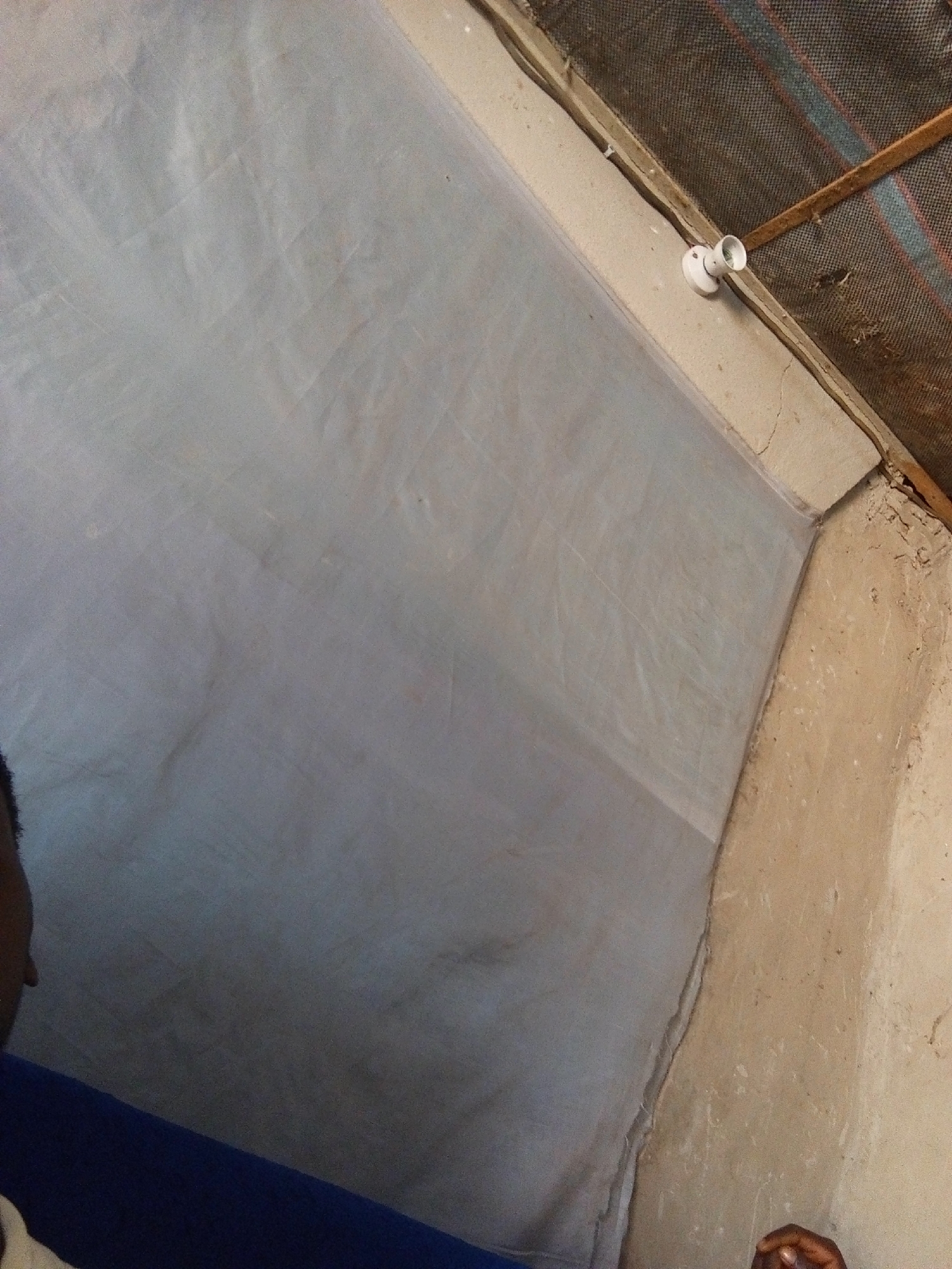 The image depicts an interior scene, likely within a structure in Nyabe, Nigeria. The foreground shows a portion of a person's head and shoulder, with dark hair and skin tone, partially visible in the lower left quadrant of the frame. This individual is wearing a dark blue garment. The majority of the image is dominated by a light gray, translucent fabric that appears to be stretched taut, covering a significant portion of the wall. This fabric is creased and folded, creating shadows and texture.
The upper right portion of the image reveals the ceiling and the junction where the ceiling meets a wall. The ceiling is constructed with wooden beams and covered by a dark mesh material, possibly for ventilation or insect protection, with visible multi-colored stripes. A simple white light fixture, a bare socket, is attached to the wooden structure near the wall. The wall itself is made of a rough, light brown, possibly mud or stucco material, showing signs of wear and small cracks. The overall impression is of a simple, perhaps temporary or utilitarian structure.