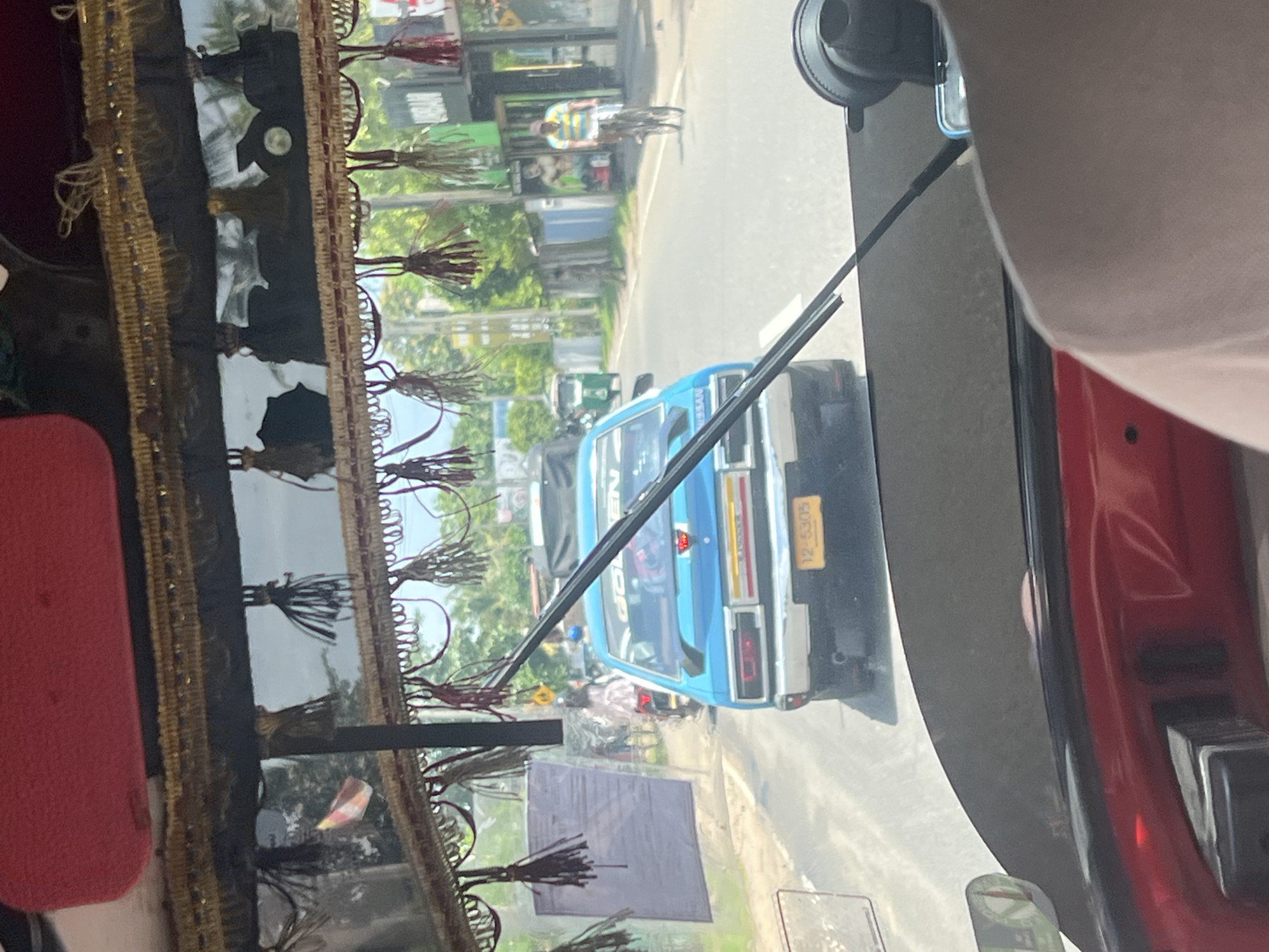 A blue sedan, possibly a Nissan, with a yellow license plate reading "12-5305" is driving on a road in Galle, Sri Lanka. The car is visible from behind, and its rear taillights and bumper are clear. The vehicle is adorned with a red and gold decorative trim hanging from the roof, with tassels. The foreground shows the interior of what appears to be a vehicle, with a red dashboard and a dark, tinted windshield. A black windshield wiper is prominent, extending across the view. To the right of the sedan, a man on a bicycle is riding down the road, wearing a yellow and blue striped shirt. In the background, buildings and trees line the street, and a green three-wheeled auto-rickshaw is also visible. The overall scene depicts a typical street view in Galle, characterized by a mix of vehicles, pedestrians, and the tropical environment.