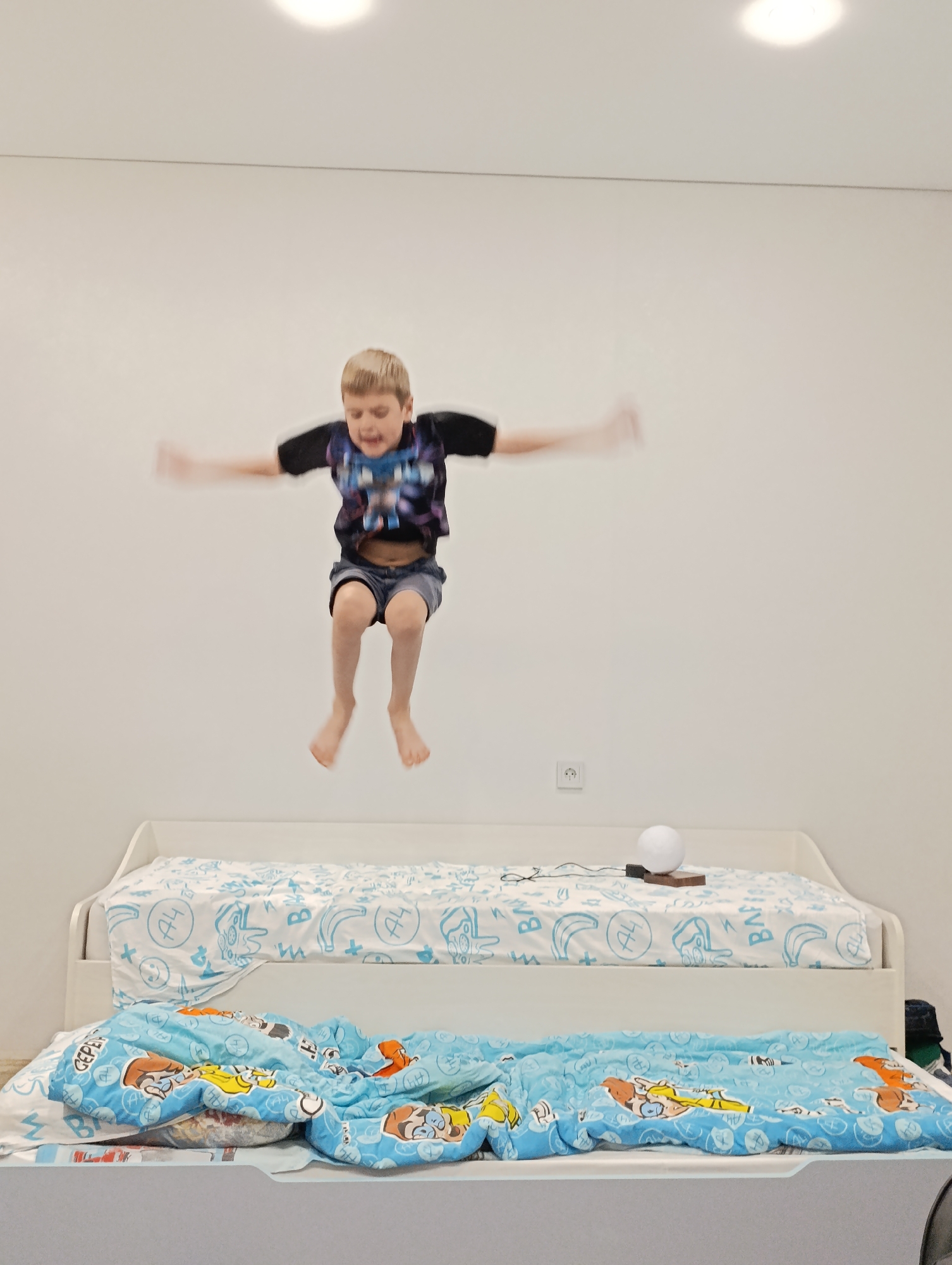 A young boy is captured mid-air, jumping on a bunk bed with blue and white patterned bedding. The boy has blonde hair and is wearing a short-sleeved t-shirt and shorts. His arms are outstretched. The bed is white, and a white spherical lamp sits on the top bunk. The background is a plain white wall with a light fixture. The location is Chaikovsky, Russia.