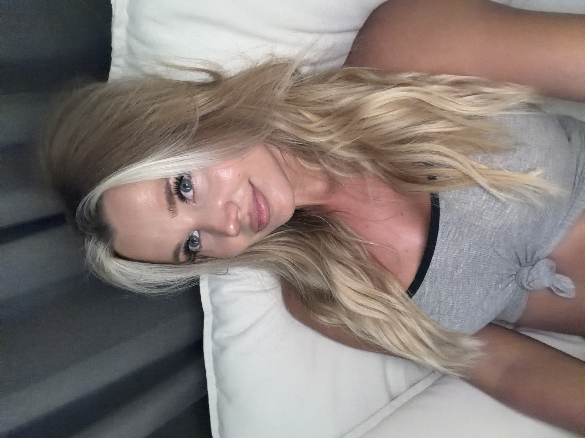 A blonde woman with blue eyes is shown from the chest up, lying on a white surface, likely a bed or sofa. She is wearing a grey tank top with a black trim and a knot in the front. Her blonde hair is styled with volume and waves, with some lighter streaks. The background is dark and appears to be a curtain. The lighting is soft, creating a warm glow on her skin. The location is not discernible from the image.