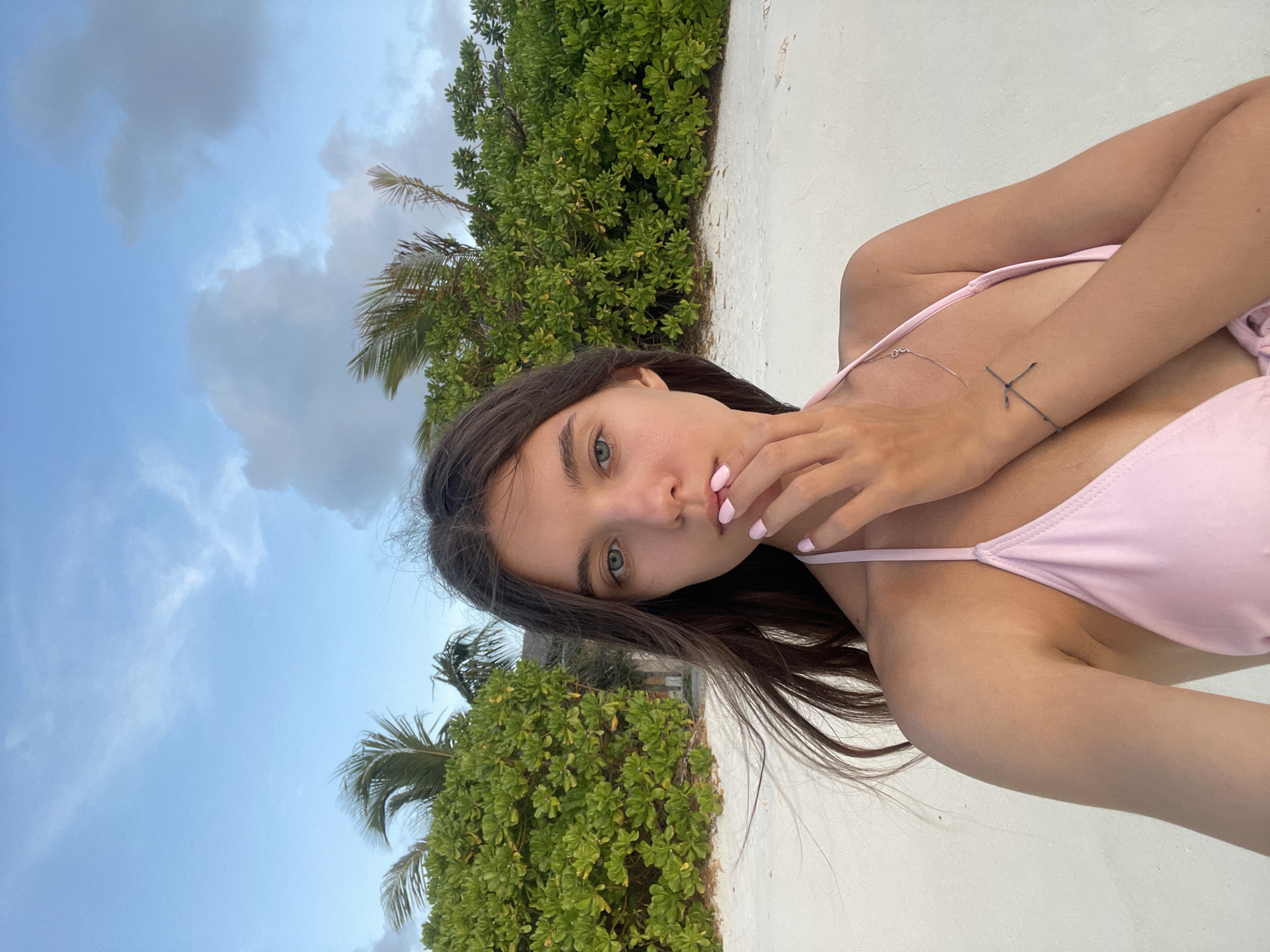 A young woman is captured in a selfie on a beach. She is wearing a light pink bikini top and has her hand on her face, obscuring part of her mouth. Her hair is dark and wavy, and her eyes are light blue. A subtle necklace and a bracelet with a cross charm are visible on her arm. The background features lush green foliage and palm trees against a cloudy sky. The ground is covered in white sand. The scene is lit by natural daylight, suggesting it is either early morning or late afternoon.