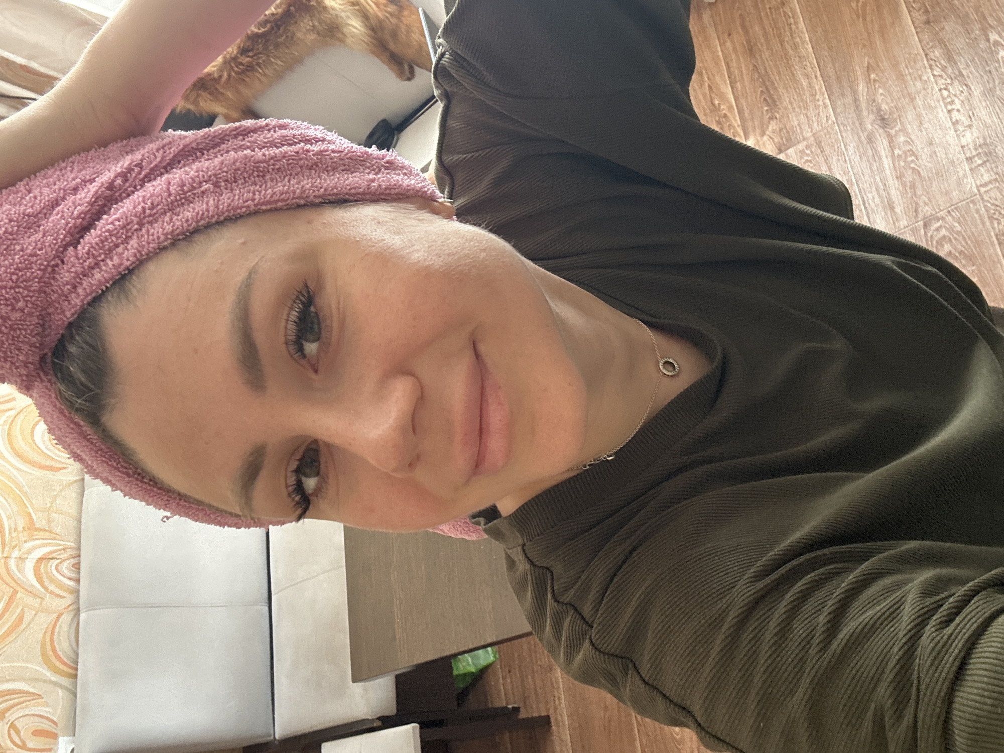 A woman is taking a selfie indoors, wearing a pink towel wrapped around her head and a dark green shirt. She has on makeup, including eyeliner and mascara. A silver necklace with a circular pendant is visible. In the background, there is a couch and a wooden floor. A cat is partially visible in the background, on a surface behind the woman. The image appears to have been taken in a residential setting, possibly an apartment in Novoaltaysk, Russia, based on the context provided.