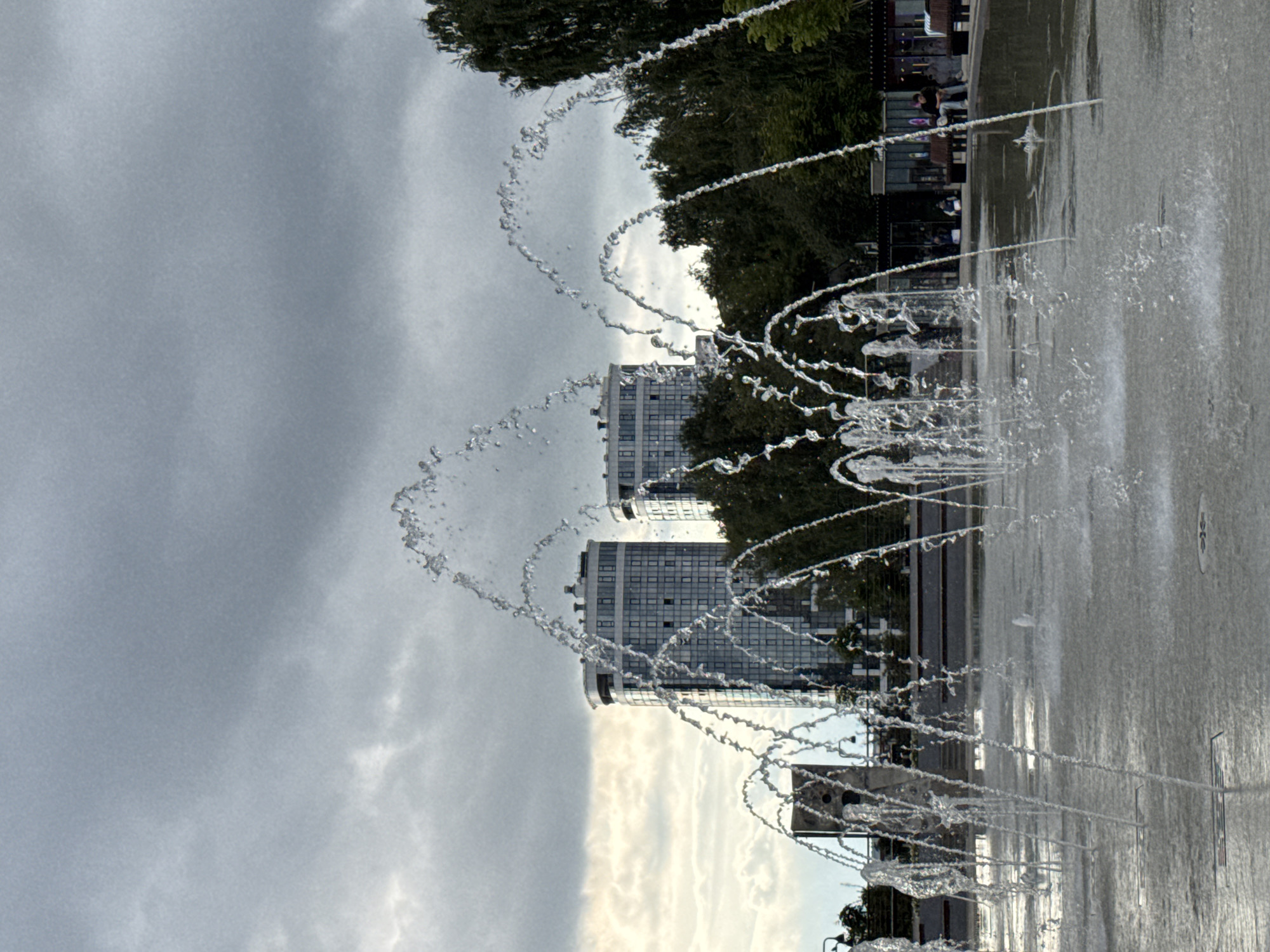 The image depicts a water fountain in a city park with two modern high-rise buildings in the background. The fountain is active, with streams of water arcing and splashing. The sky is overcast with gray clouds. In the background, there are trees and a few people sitting on benches. The scene is set in a public urban space, likely a park or plaza, with the architecture suggesting a contemporary city environment. This location is identified as being in Izhevsk, Russia.