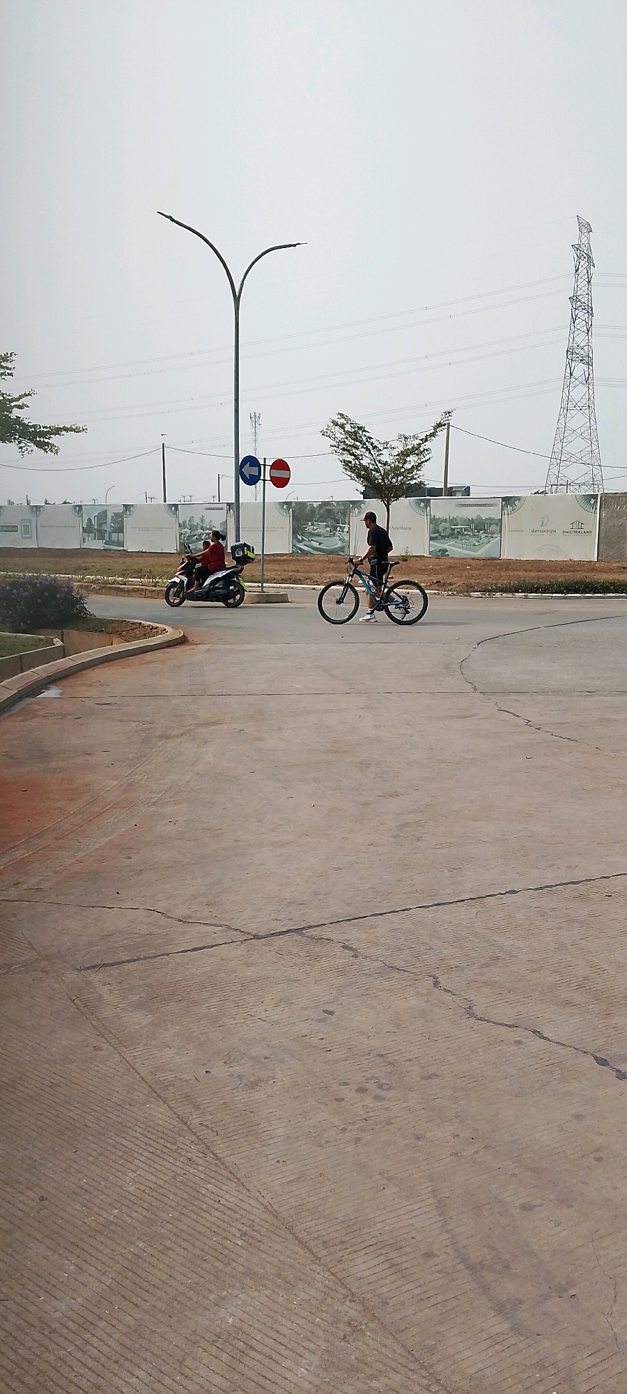 In Satriamekar, Indonesia, on a hazy day, a concrete road curves in the foreground. To the left, a motorcycle carrying two individuals, one wearing a red shirt, travels away from the camera. To the right, a person wearing a black t-shirt and shorts pushes a blue bicycle. In the background, a large billboard displays renderings of residential buildings, suggesting a development area. Beyond the billboard, power lines stretch across the sky, supported by a tall transmission tower on the right and a street lamp with a curved pole on the left. Traffic signs, including a blue arrow indicating a left turn and a red and white circular sign, are visible near the center. The environment appears to be a developing urban or suburban landscape.