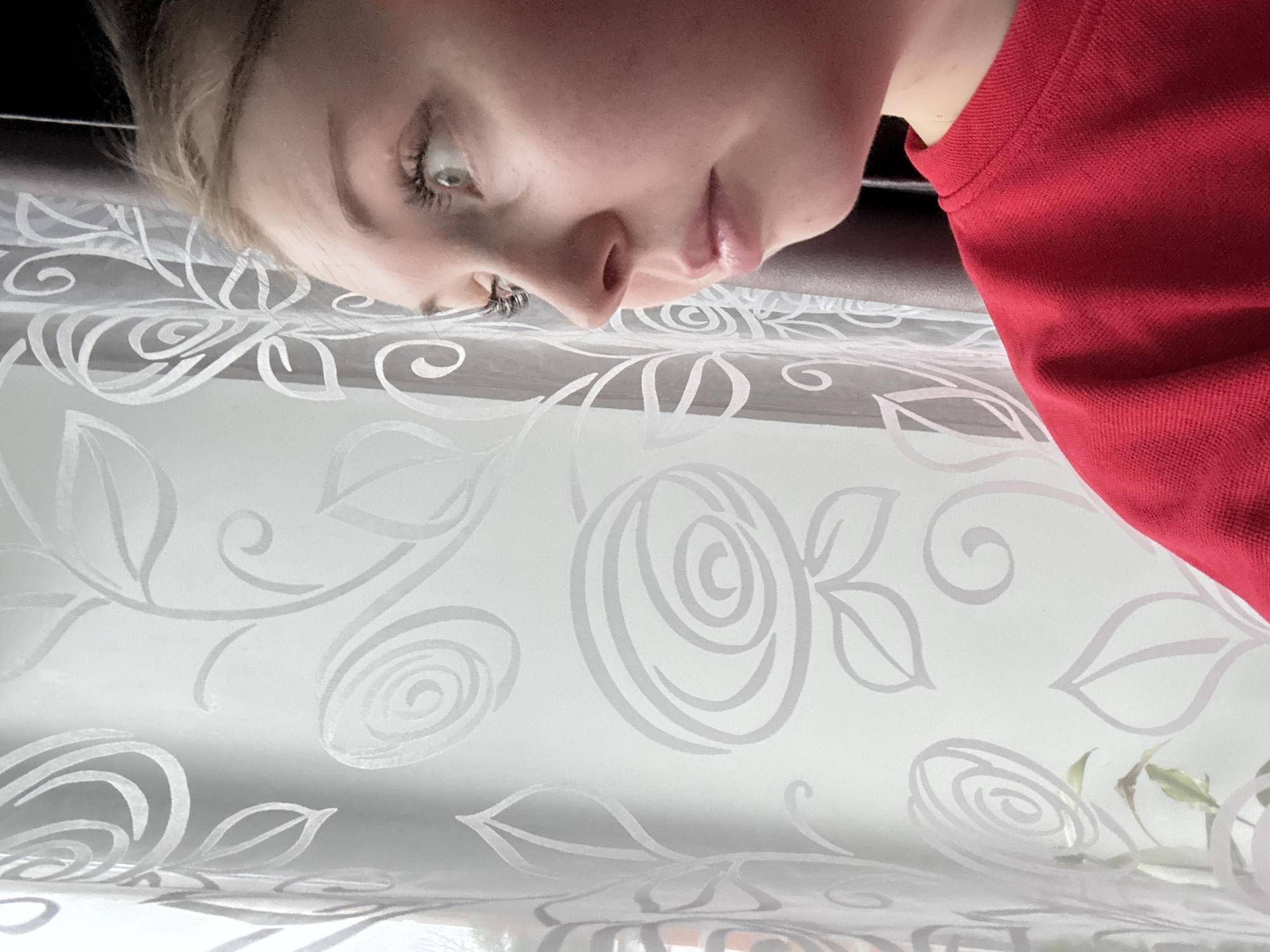 The image presents a close-up, inverted view of a person's upper body and face against a background of patterned fabric. The subject, likely female, has fair skin, light hair, and light-colored eyes. Her eye, with visible eyelashes, is prominent on the left, and her nose and slightly parted lips are visible towards the center. She appears to be wearing a red garment, visible on the right side of the frame.
The majority of the image is filled by a sheer, white fabric, likely a curtain or drape, featuring a repeating pattern of stylized gray roses and leaves. This pattern is oriented upright relative to the bottom of the frame, indicating that the person is seen from an inverted perspective. The lighting suggests a bright, possibly daytime, setting. The person's expression is neutral and contemplative, with her gaze directed downwards towards the fabric. There is no specific activity taking place other than observation. No text is visible in the image.