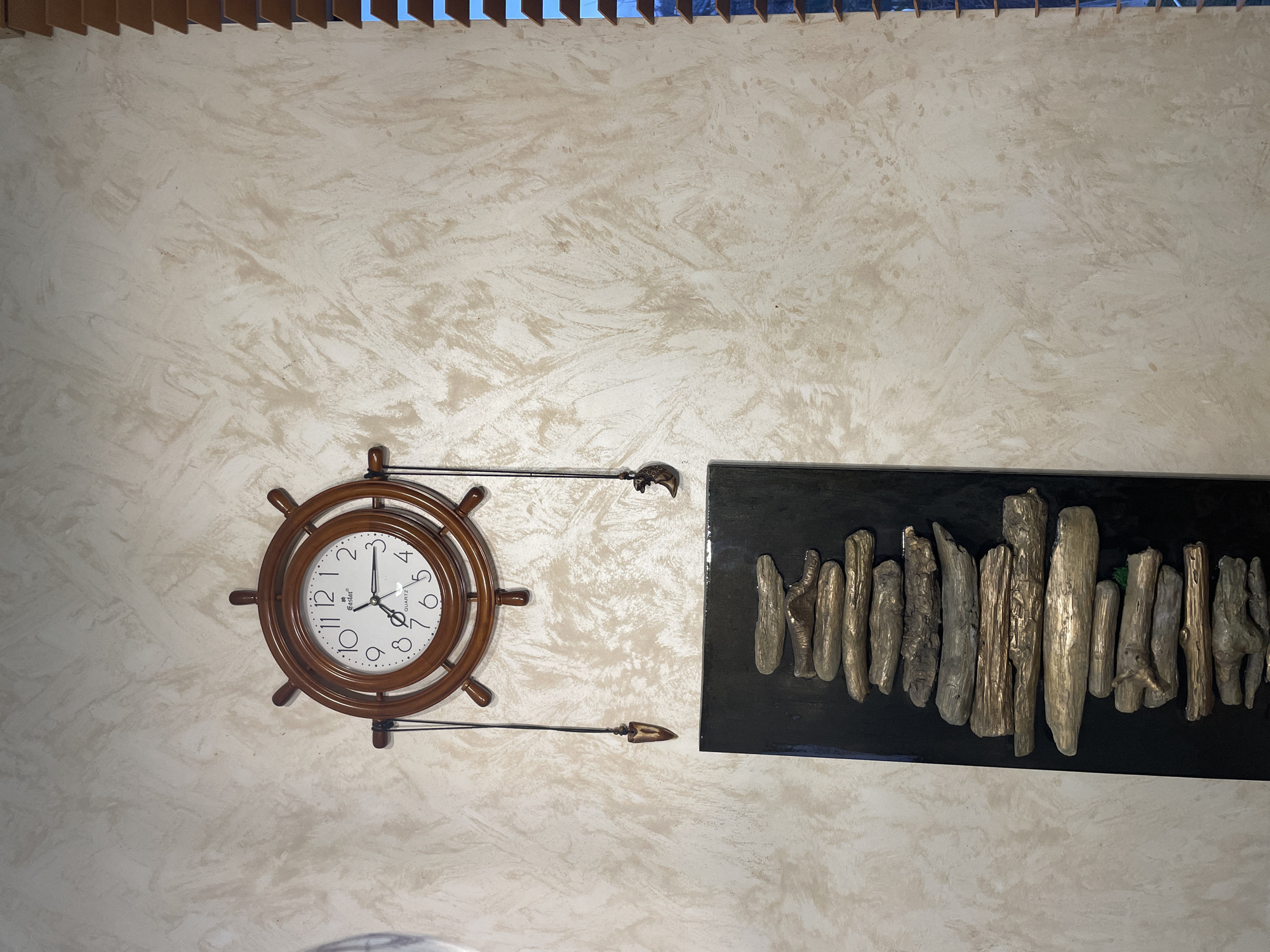 This image shows a wall with a decorative clock and an artwork in what appears to be an interior setting. The clock is designed to resemble a ship's wheel, with a brown wooden frame and black hands. The artwork to the right is a black rectangular piece featuring vertically arranged driftwood or similar objects of varying sizes, possibly adhered to the backing. The wall itself is textured, with a light beige or off-white color. There are no people visible in the image. The presence of Russian letters on the clock face possibly confirms it's location in Лесной Пейзаж Кп (шарапово), Russia.