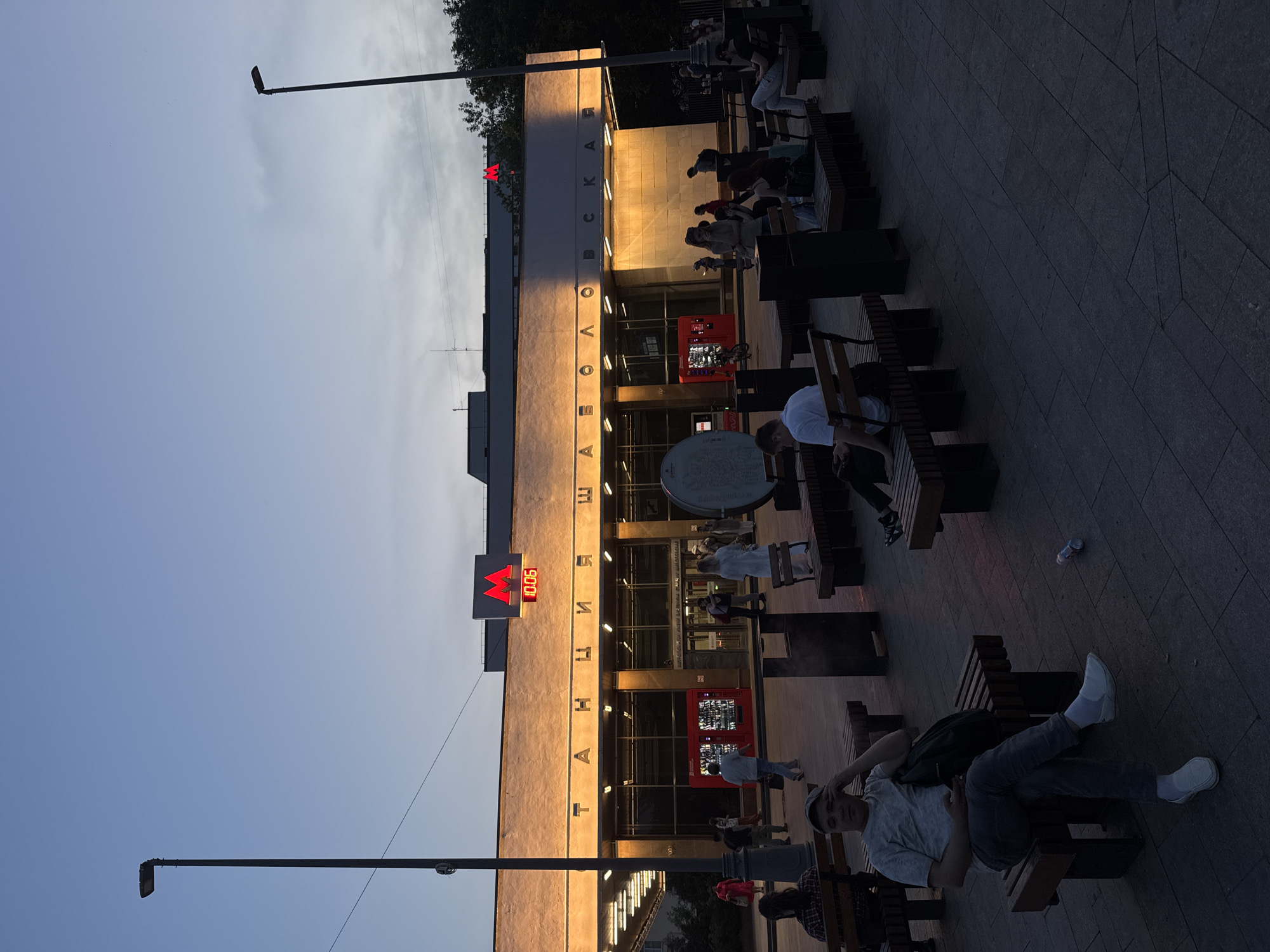 The image depicts the exterior of the Shabolovskaya metro station in Moscow, Russia, at dusk. The station's facade is illuminated by warm, artificial lights, highlighting the prominent red "M" logo of the Moscow Metro and the station's name, "СТАНЦИЯ ШАБОЛОВСКАЯ", in Cyrillic lettering. A digital clock displays "10:06".
In the foreground and midground, several individuals are seated on benches. A man in a grey t-shirt and dark pants is seated on a bench on the left, leaning his head on his hand, appearing to be looking at his phone. A bottle lies on the ground near him. Other people are visible on benches further back, some facing the station, others facing away. Some are interacting with each other, while others appear to be alone.
The overall environment suggests a public space outside the metro station, with paved ground and trees visible in the background. The sky is a deep blue, indicating the transition from day to night. Two tall streetlights are also prominent features of the scene. The lighting creates long shadows across the paved area.
