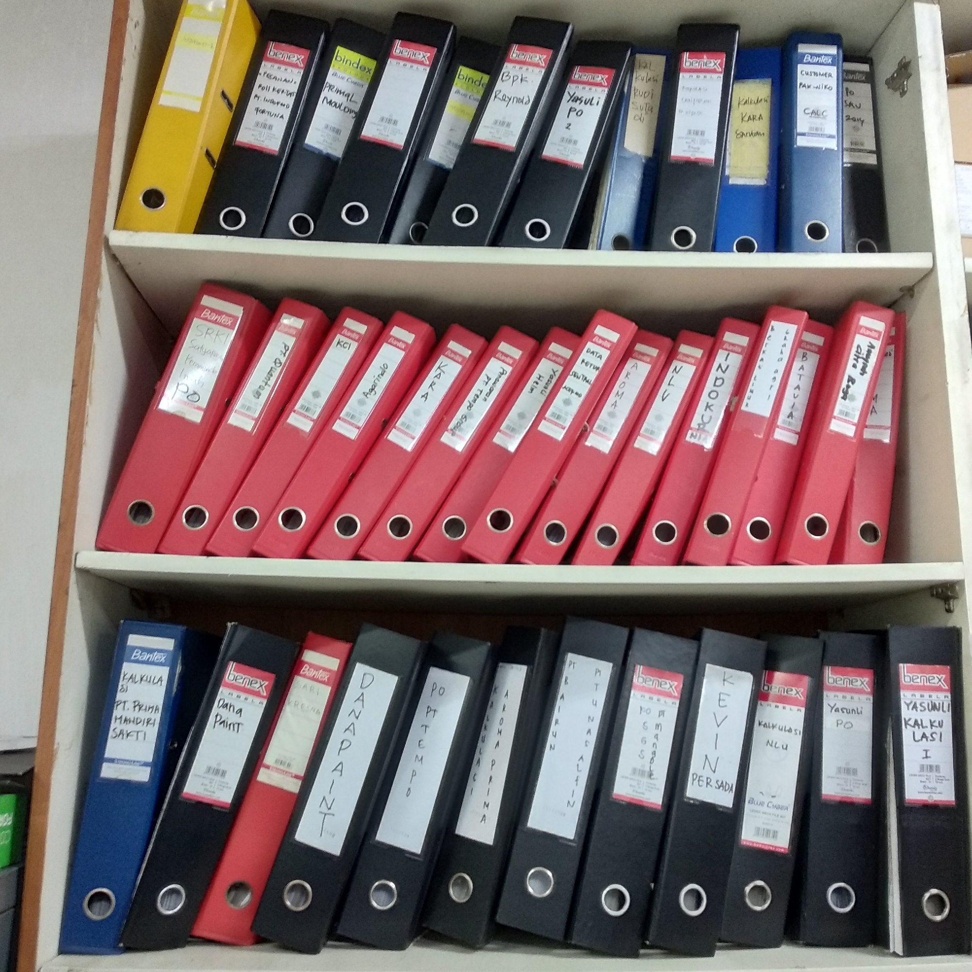 This image shows a three-tiered bookshelf filled with binders, likely in an office setting. The binders are of various colors, including yellow, black, blue, and red, and some have labels with handwritten text. The bookshelf itself appears to be made of wood and is positioned against a wall. The overall environment suggests an organized storage space, possibly for documents or files.
The location in Balaraja, Indonesia, indicates a setting within that specific city. The contents of the binders, such as labels with Indonesian names and terms like "PT" (Perseroan Terbatas, a form of limited liability company in Indonesia), strongly support this geographical context. The presence of multiple binders filled with what appears to be administrative or financial documentation suggests that this is a workplace or an office environment where record-keeping is essential.
The image does not contain any people or visible actions or interactions. The focus is solely on the arrangement and contents of the binders on the bookshelf. The scene is static and conveys a sense of order and routine.