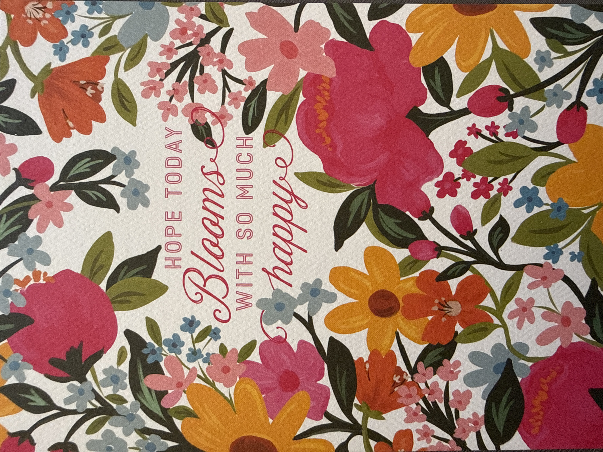 The image is a close-up shot of a greeting card with a floral design. The card features a white background with an abundance of colorful flowers, including pink, orange, yellow, and blue blossoms, interspersed with green leaves and dark stems. The text on the card is in a decorative font and reads, "HOPE TODAY Blooms WITH SO MUCH happy". The flowers are depicted in a stylized, painterly manner. There is no discernible location in the city of West Sacramento, United States of America, as the image is of an indoor item.