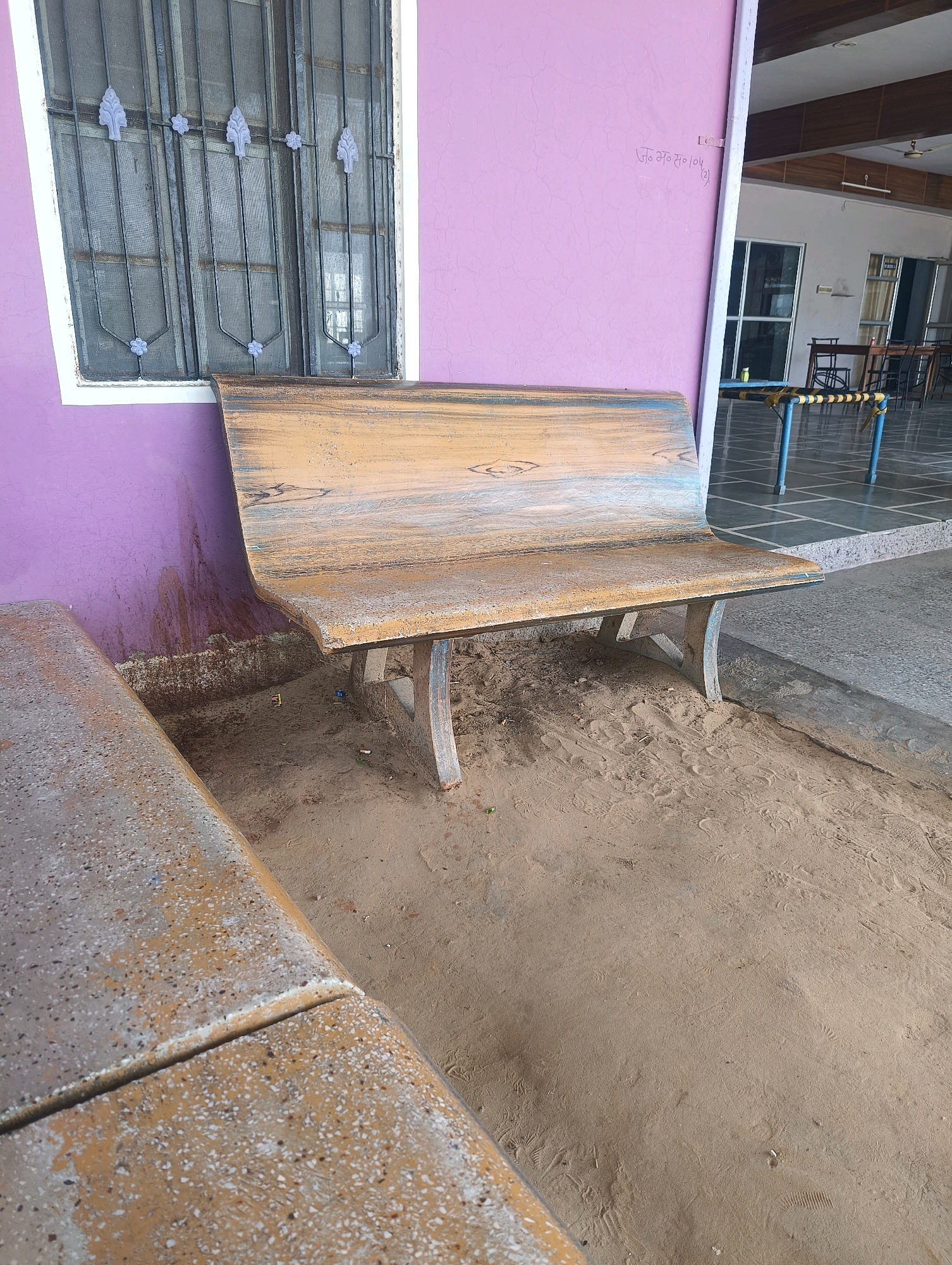 In Chhapri Bari, India, a long, curved, concrete bench with a wood-grain-like finish is situated outdoors on sandy ground. The bench is positioned in front of a purple-painted wall with a window that has decorative iron bars and mesh screens. To the left of the main bench, another concrete bench, with a speckled finish, is partially visible. In the background, there is a tiled floor with a raised platform, suggesting an indoor or semi-outdoor seating area with tables and chairs. The ground around the benches is sandy with visible footprints and some scattered debris, including cigarette butts. The overall scene depicts an outdoor seating area that appears to be part of a public or commercial establishment.