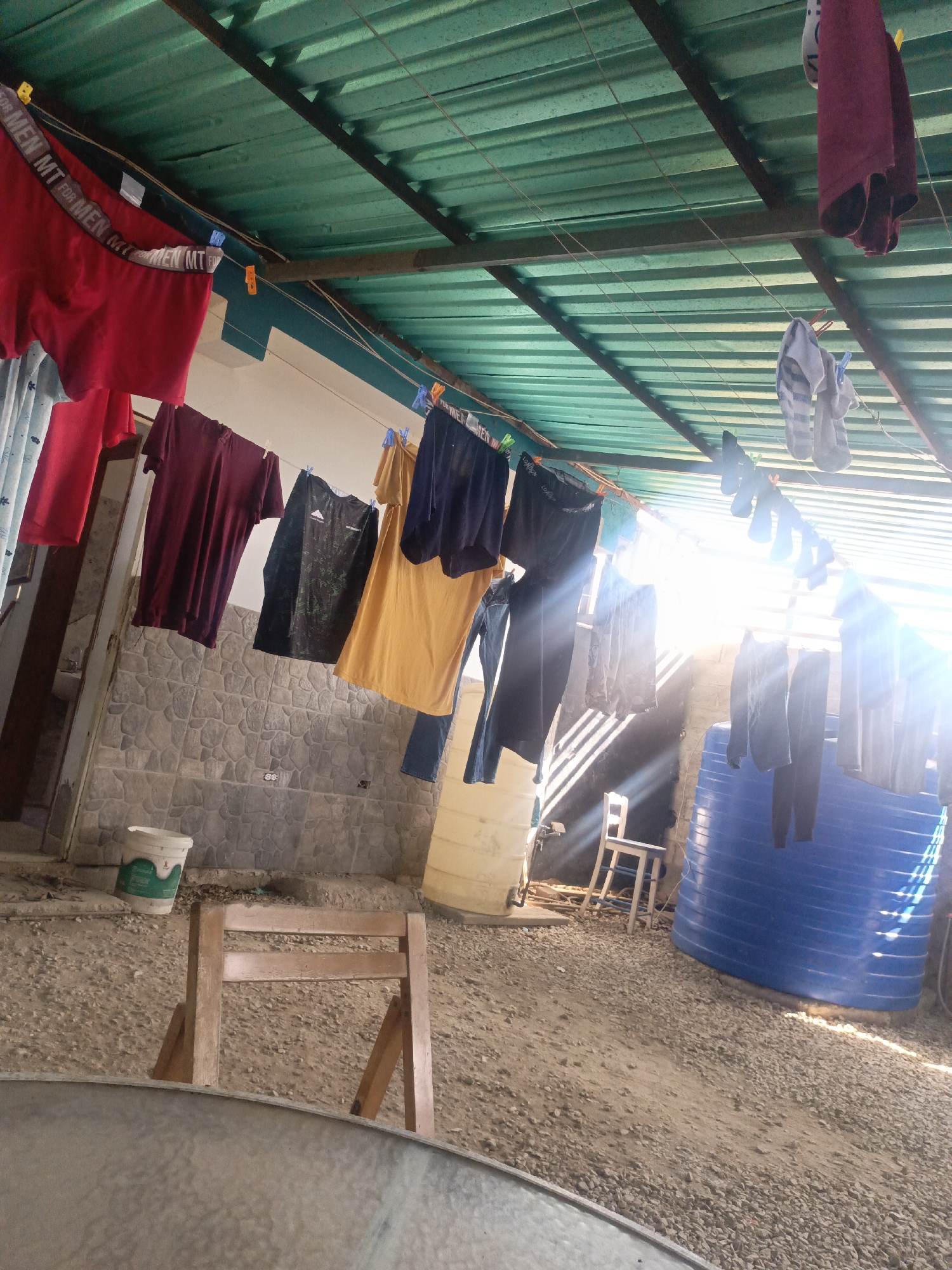 The image depicts an outdoor space, possibly a patio or yard, with a corrugated green roof. Laundry is hung on a clothesline strung beneath the roof. The laundry consists of various garments, including shirts, pants, underwear, and socks. The clothes are secured to the line with clothespins.
In the background, there is a light-colored stone wall, along with a large white storage container and a blue water tank. A simple wooden chair is visible behind the clothesline. The ground is covered with a gravel or dirt surface.
The image is brightly lit, with strong sunlight streaming in from the right side of the frame. A part of a table is visible in the foreground. The location is likely Juan Griego, Venezuela, based on the instructions, although no explicit features in the image confirm the city.