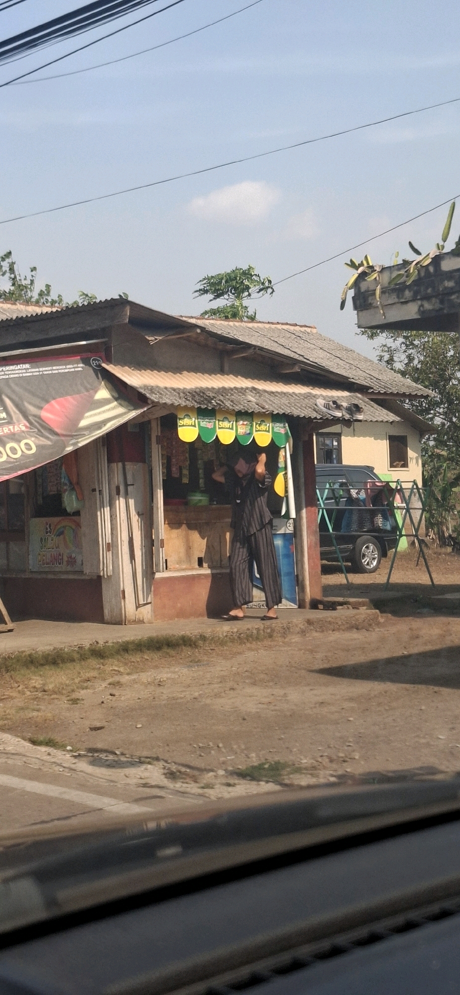 The image shows an outdoor scene in Cibinong Hilir, Indonesia, on a sunny day. In the foreground, a portion of a car's dashboard is visible, reflecting the surroundings. The main focus is a small roadside stall with a corrugated metal roof. A person wearing a dark striped outfit is standing behind the counter, appearing to be arranging items. The stall is stocked with various goods, including what appear to be food items and drinks displayed on hangers. A banner with Indonesian text and an image of a red object is partially visible on the left side of the stall. Behind the stall, a dark-colored vehicle is parked. The ground is a mixture of dirt and gravel, with some patches of grass. Electrical wires are strung across the sky, and a few trees are present in the background.