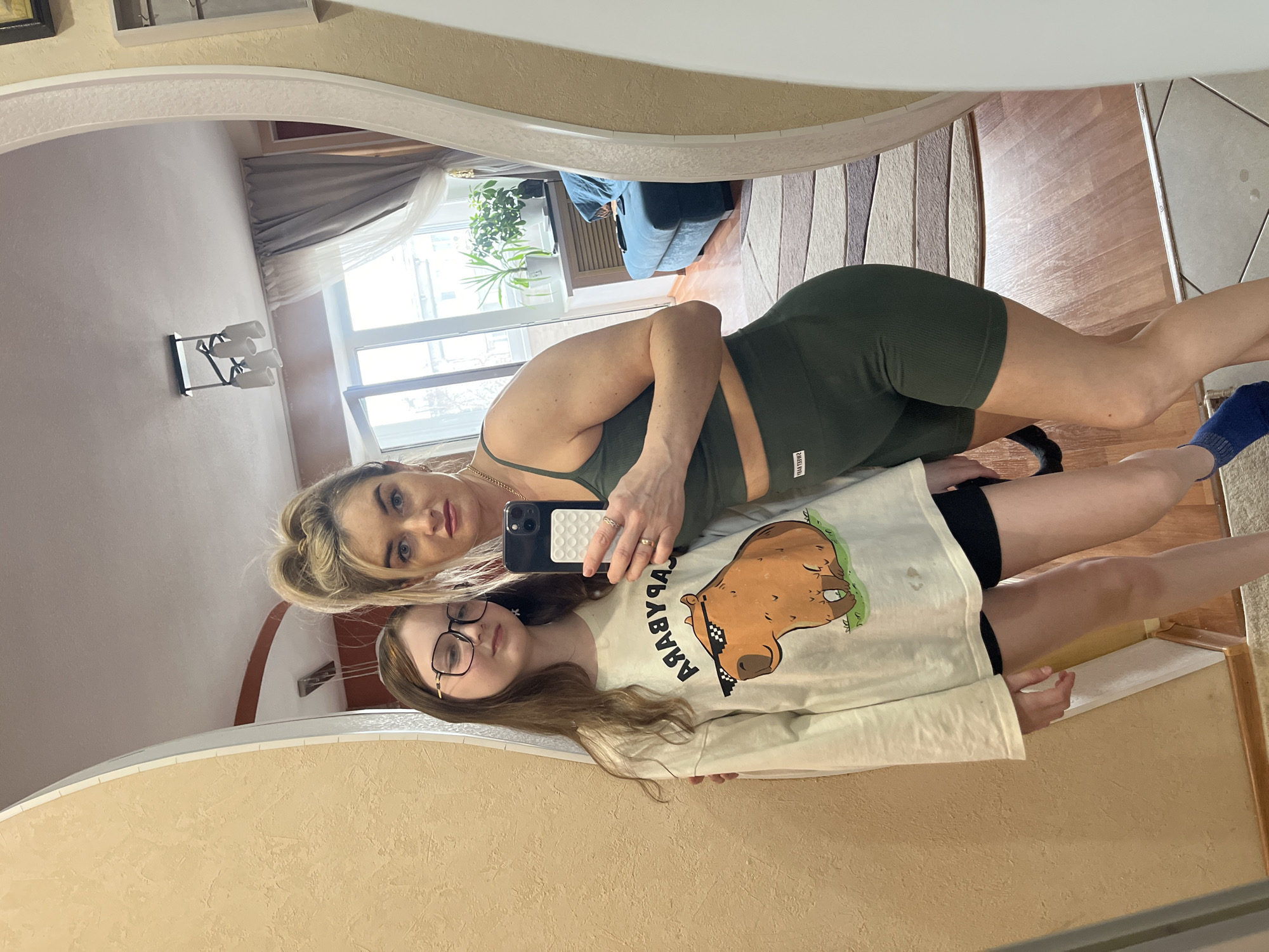 Two females are posing for a selfie in a mirrored reflection. The older female has blonde hair pulled back in a bun and is wearing a green athletic top and matching shorts. She is holding a black iPhone in her hand. The younger female is wearing glasses and a long-sleeved white shirt with a capybara cartoon printed on it, along with black shorts. The reflection shows a room with a large window, curtains, and a glimpse of furniture. The location is Chaikovsky, Russia.