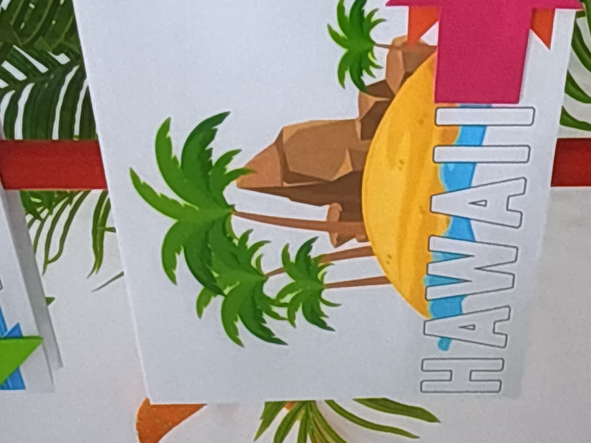 The image shows a close-up of a sign with the word "HAWAII" written in large, outlined white letters against a backdrop of a cartoonish island scene. The island features a yellow sandy beach, blue water, brown rock formations, and several green palm trees. A bright red pole is visible in the background, suggesting the sign is part of a larger display. The scene is colorful and illustrative, with a tropical theme. There are no people or animals present in the image. The location is not specified as being in Dubai, United Arab Emirates; the sign itself depicts Hawaii.