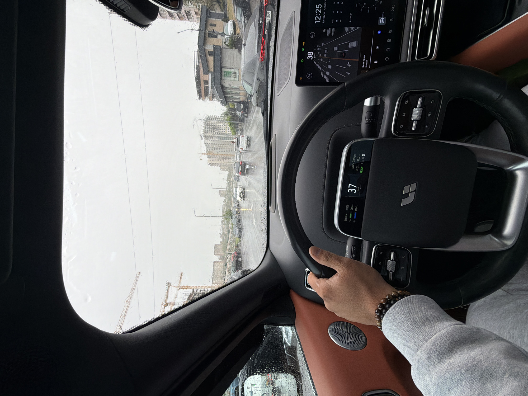 The image shows the interior of a car driving on a rainy day. A hand wearing a bracelet is on the steering wheel. The dashboard displays information like speed (37 km/h) and battery status, along with a navigation screen. Outside, a city landscape is visible, including buildings, construction cranes, and other vehicles. The dashboard screen displays the time, "12:25, Sat May 16 Cloudy" indicating the date and weather conditions. The navigation screen displays the words "Были танцы БЬЯНКА" and "38". A license plate of a vehicle in front reads "B 349". While specific buildings cannot be determined from the image to identify Yerevan precisely, the presence of construction cranes and a mix of older and newer buildings suggests an urban environment with ongoing development.