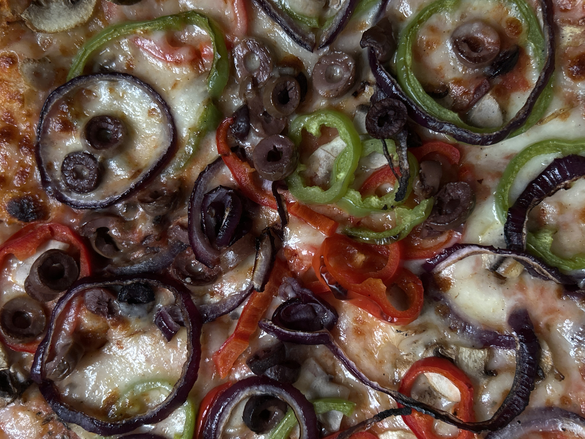 This is a detailed, top-down close-up shot of a baked pizza, focusing on its surface and toppings. The pizza is generously covered with melted, bubbly cheese, which has some golden-brown, slightly crisped areas. Various colorful vegetables are visible as toppings. These include prominent rings of purple-red onion, slices of bright green bell pepper, and slices of red bell pepper. Black olives, typically sliced into rings, are scattered throughout the pizza. Light brown mushroom slices are also discernible beneath the cheese and other toppings.
The lighting appears soft and even, highlighting the glossy texture of the melted cheese and the distinct colors and shapes of the vegetables. The scene captures the fresh, cooked appearance of the ingredients. There are no people, discernible activities, or visible text in the image. The image is a still-life food shot, likely taken indoors. While the context is Sofia, Bulgaria, this information is not visually identifiable from the pizza itself.