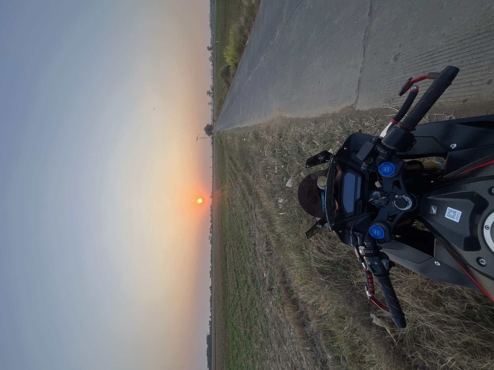 An outdoor, eye-level, long shot captures a motorcycle parked on the side of a paved road next to a field. The sun is setting in the distance, casting an orange glow across the sky and the field. The motorcycle is black and red, with the handlebars and front wheel visible in the foreground. The field is mostly dry grass, with some green patches. A paved road stretches into the distance, with a single street lamp visible on the side. The scene appears to be in Troso, Indonesia, based on the context provided.