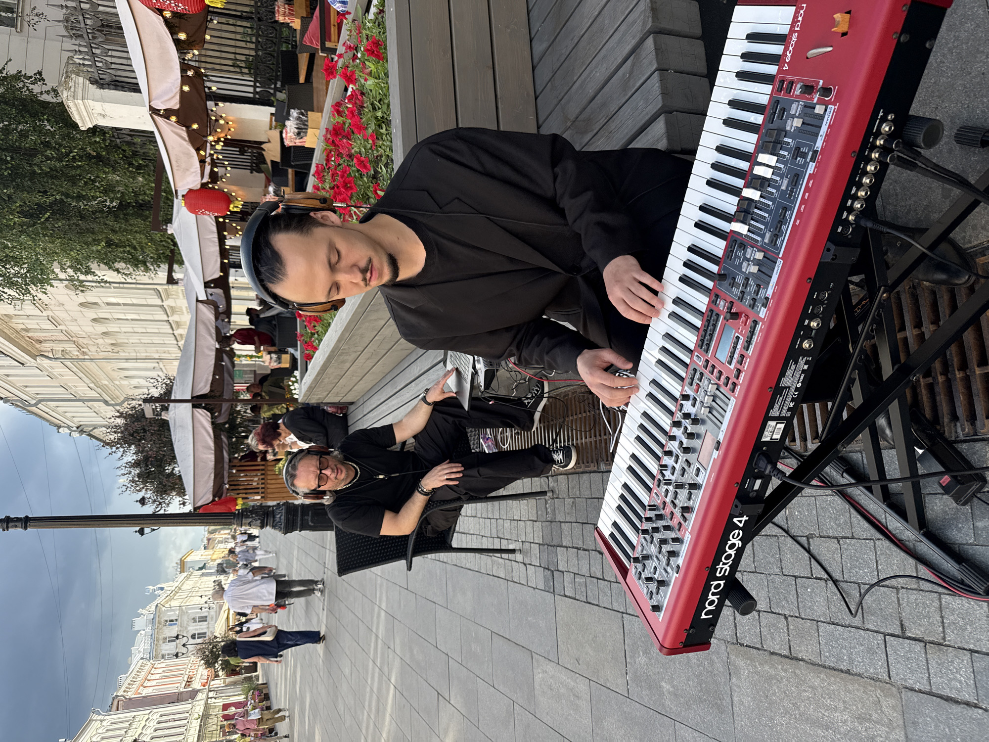 The image depicts an outdoor scene, likely a street or pedestrian area, with people and a musician. In the foreground, a man wearing headphones and a black outfit is seated, playing a red Nord Stage 4 keyboard. He appears focused on his music, with his eyes closed. Another person, also in black, is seated nearby, possibly assisting or accompanying the musician. The keyboard is set up on a stand, with cables and equipment visible.
The background reveals a street lined with buildings, some with awnings and umbrellas, suggesting a commercial or cafe area. There are other people in the distance, some walking and others seated at tables, indicating a public gathering or a leisurely day. Red flowers are blooming in a planter box near the musician. The time of day appears to be daytime, with soft lighting. The overall mood seems relaxed and somewhat artistic, with the musician performing in a public space. Visible text on the keyboard reads "nord stage 4". The location is identified as Nizhny Novgorod, Russia.