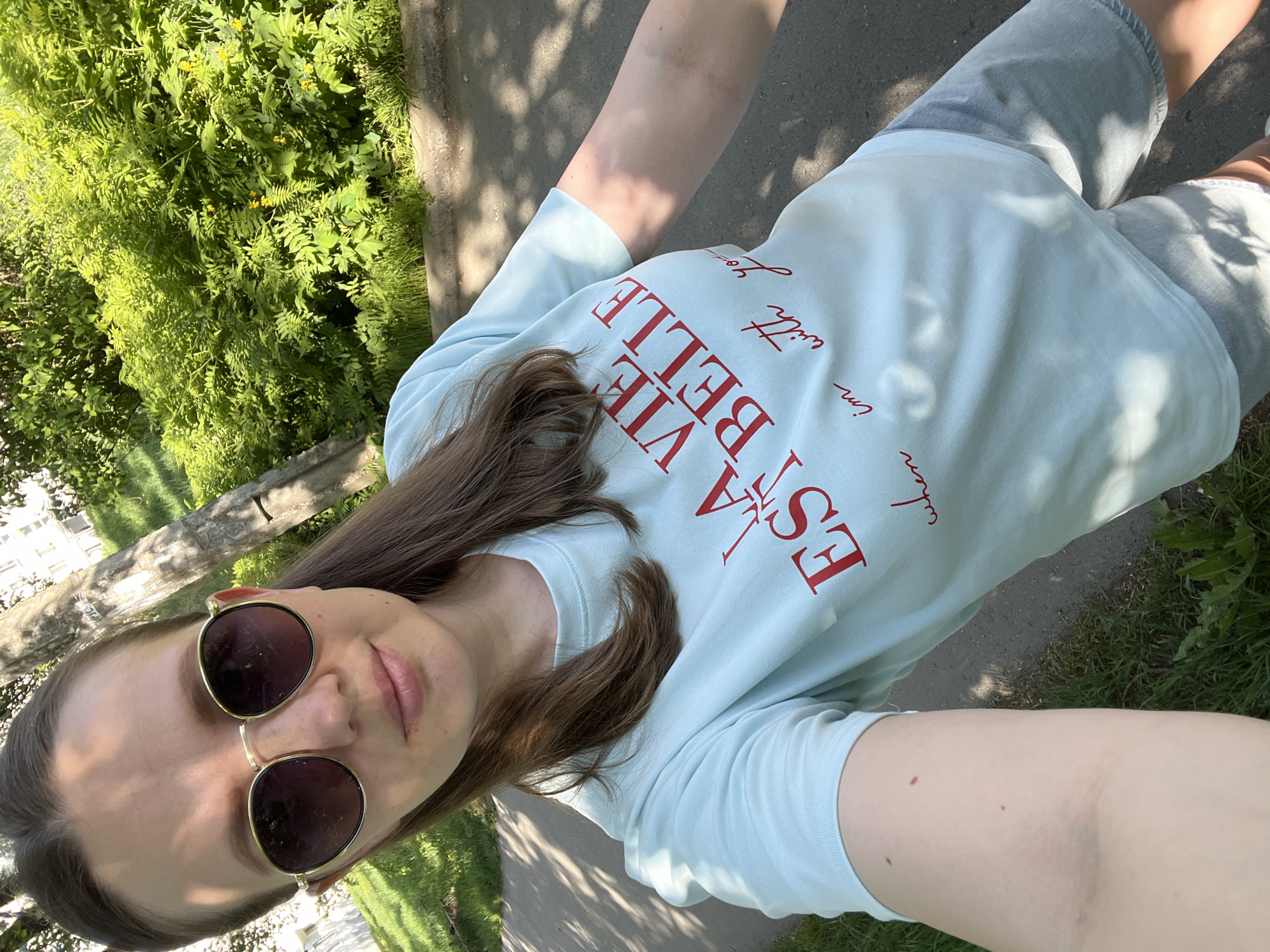 A selfie taken on a sunny day in Chaikovsky, Russia. The individual, wearing round sunglasses, a light blue t-shirt with red lettering, and gray shorts, appears to be standing on a paved pathway alongside green bushes and grass. A building is visible in the background to the left. Shadows from trees are cast on the ground.