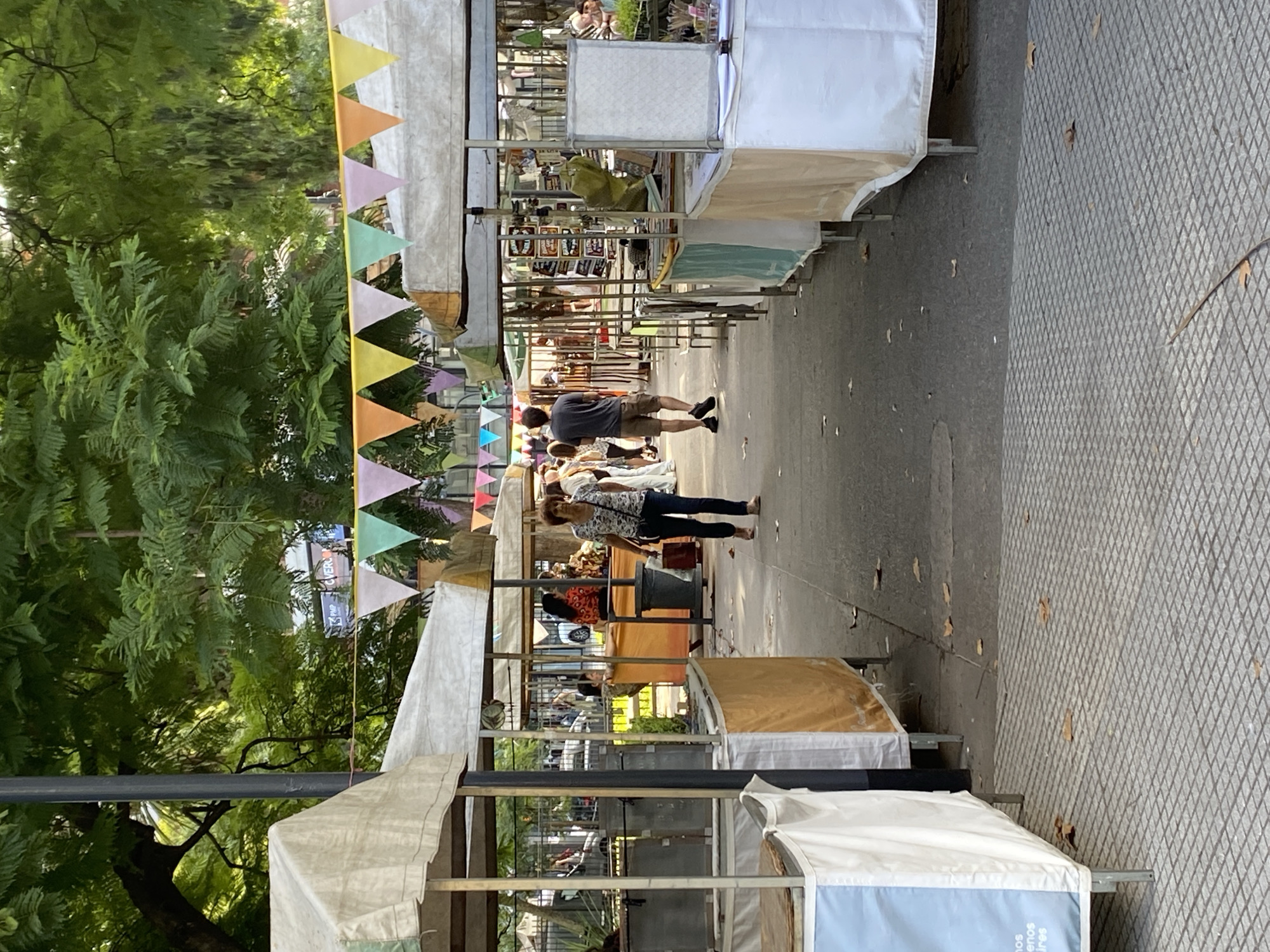 This image shows a row of vendor stalls set up along a pedestrian walkway, likely in an outdoor market in Buenos Aires, Argentina. The stalls are covered by white canopies, and some have colorful triangular flags hanging above them. People are walking through the market, browsing the goods on display. Trees line the walkway on the left side of the image, providing shade. The ground is paved with patterned stones or tiles.