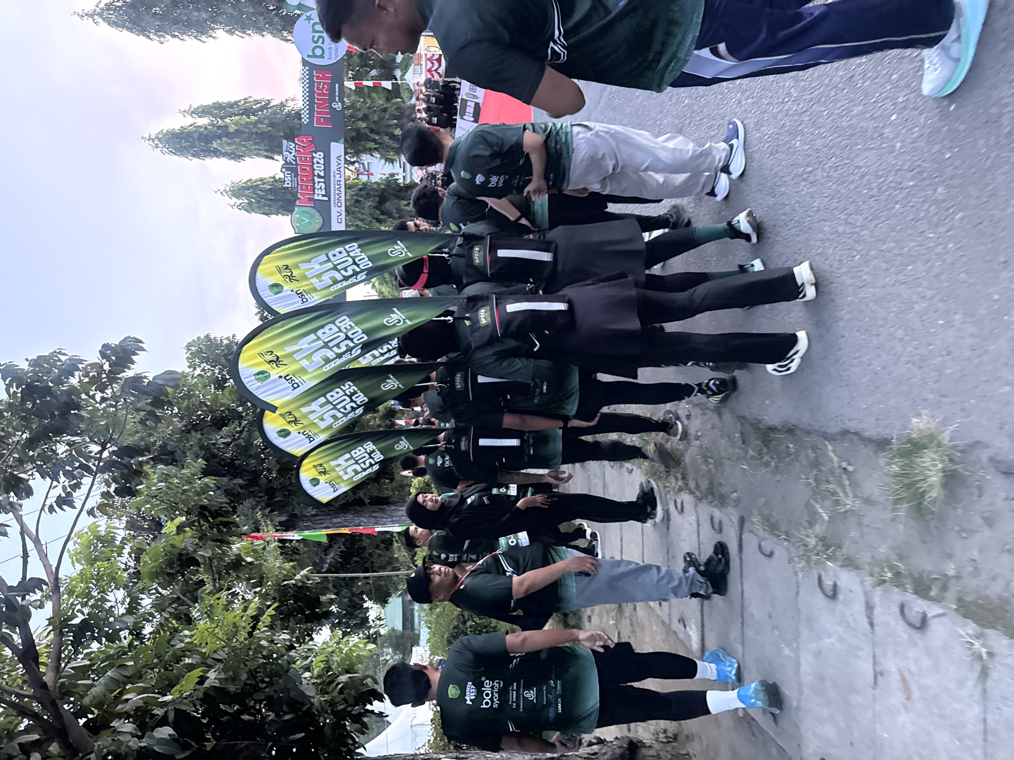 The image depicts an outdoor event, likely a 5K race, in Lhokseumawe, Indonesia. A group of participants, predominantly wearing dark green shirts with event branding, are gathered on a paved pathway lined with grass and trees. Some individuals are wearing race bibs and backpacks. In the background, a large banner announces "MERDEKA FEST 2026" and "FINISH," with additional sponsor logos. The sky suggests either early morning or late afternoon lighting. The overall scene conveys a sense of organized community activity within a city setting.