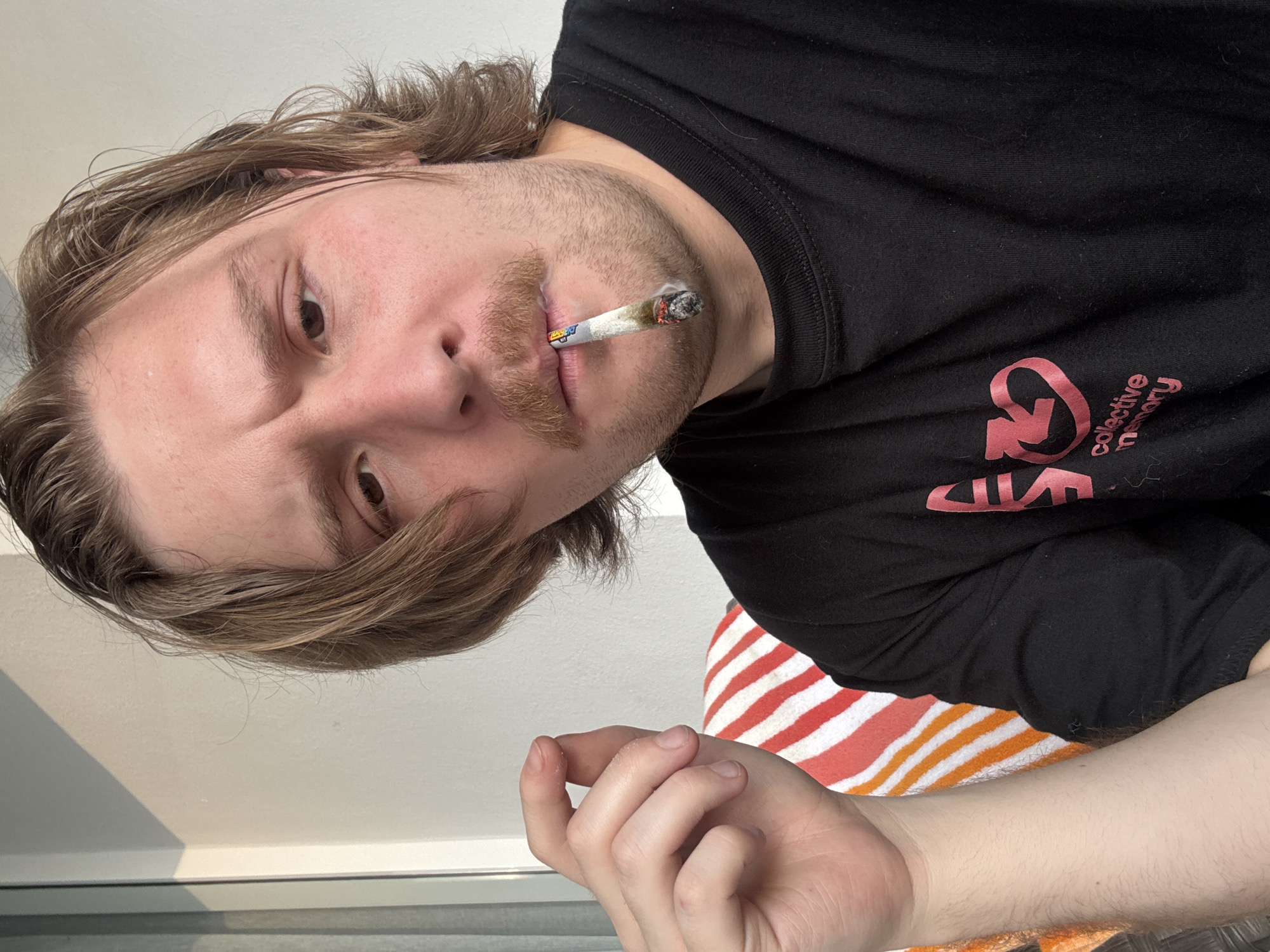 A fair-skinned individual with long, light brown hair and a mustache is shown from a close, low-angle perspective. They are holding a lit cigarette in their mouth. They are wearing a black t-shirt with a pink graphic on it. A striped orange, red, and white fabric is visible in the background. The photo appears to be taken indoors against a white wall. There is no information to suggest that the photo was taken in the city of Huixquilucan de Degollado, Mexico.