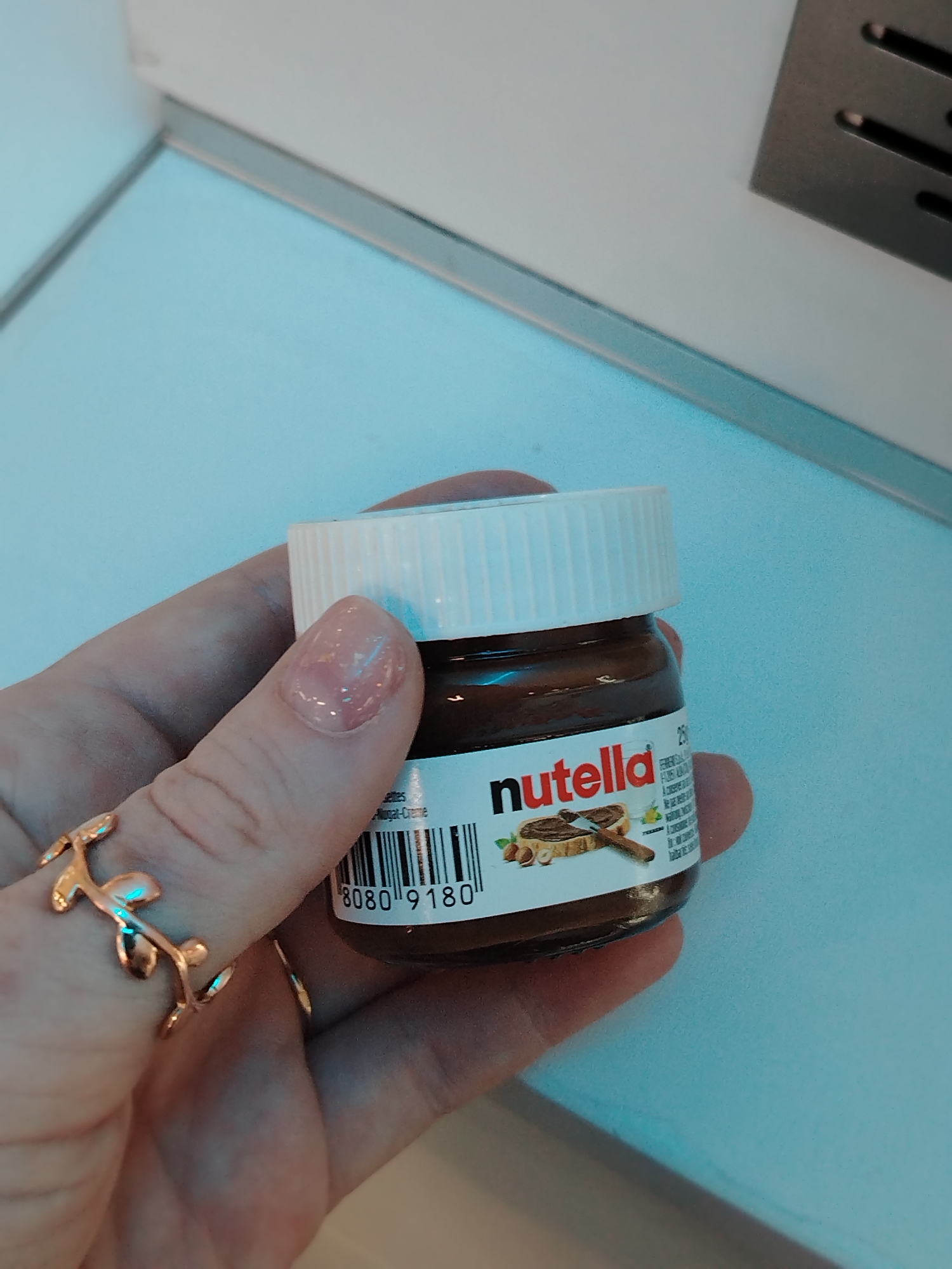 A person's hand, adorned with a gold-colored ring shaped like a twig with leaves, holds a small jar of Nutella. The jar has a white lid and a label with the "Nutella" logo prominently displayed. A barcode is also visible on the label, reading "8080 9180". The background is a light blue surface with a white edge along the top, and a metallic, slotted vent is visible in the upper right corner. There is no information available from the image about the city of Izhevsk, Russia.
