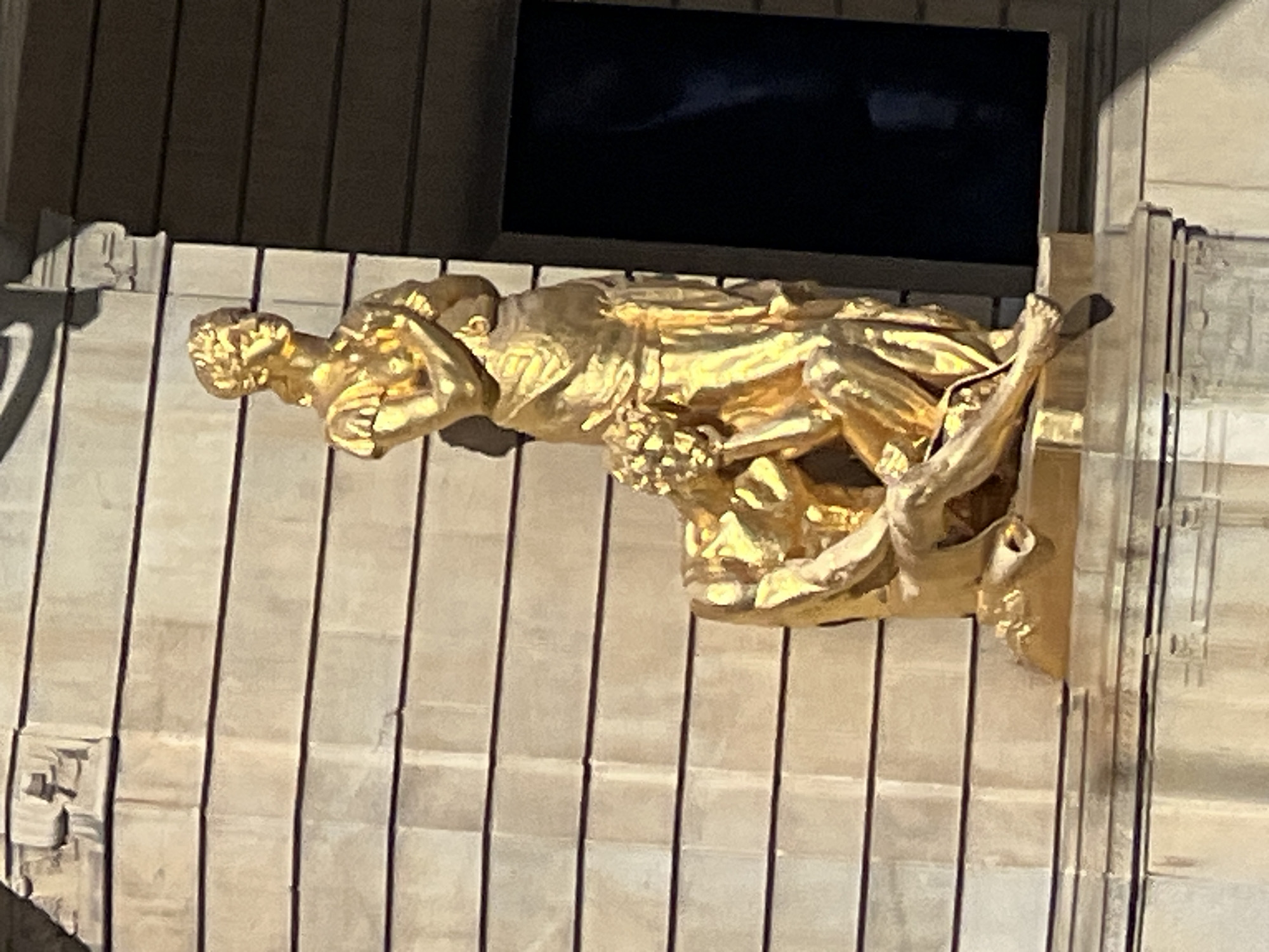 The image depicts a golden sculpture featuring two figures in front of a building with a facade of horizontally layered stone blocks. The sculpture is positioned on a ledge above a gray stone base. In the foreground, a male figure is depicted kneeling, with his head bowed and hands resting on his knees. He is wearing a short tunic. Behind him stands a female figure, also in golden attire, with her arms crossed over her chest and her head tilted slightly. She appears to be wearing a long gown with a laurel wreath on her head. The sculpture is bathed in sunlight, casting shadows on the figures and the building. To the right of the sculpture, a dark rectangular window is visible, reflecting a muted blue tone. The building facade exhibits some staining and wear, particularly on the upper left section. The overall impression is of a classical or allegorical representation. The sculpture is located on the exterior of the Swedish National Bank (Sveriges Riksbank) in Stockholm, Sweden.
