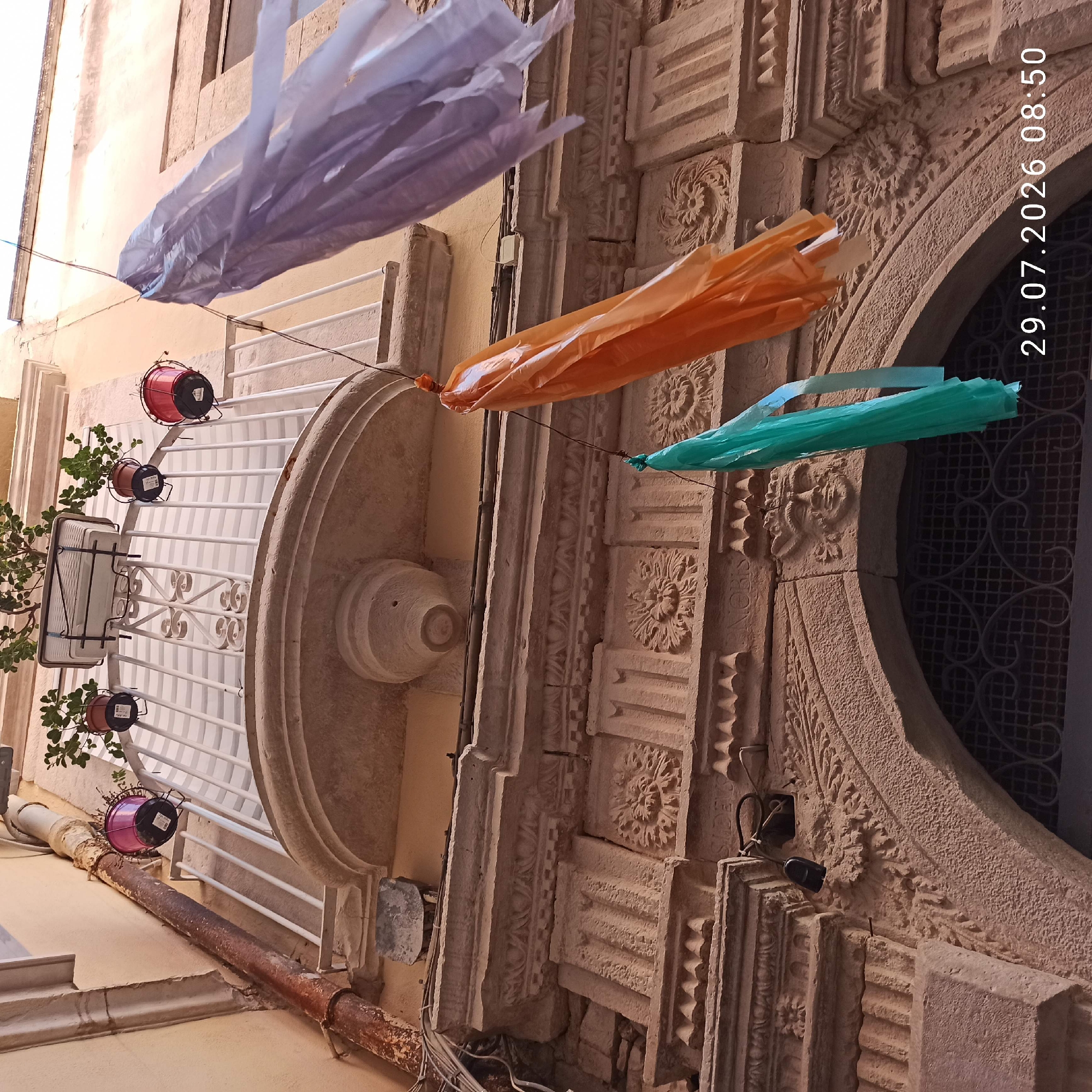 An eye-level, low-angle shot shows a view looking up at the facade of a building in Bitonto, Italy, with decorative streamers hanging from wires. Three hanging streamers, colored lavender, orange, and teal, are visible. The lavender streamer is on the right, partially obscuring the building's wall. The orange streamer is in the center, and the teal streamer is on the left, closer to the bottom of the frame. The streamers appear to be made of thin, crinkled plastic or paper.
Above the streamers, a white balcony railing with decorative metalwork is visible, with several potted plants on top. Three small, round planters are hung from the railing, one on the left with a pink pot and two on the right with red pots. A larger rectangular planter is in the center.
The building facade below the balcony features intricate stone carvings. A decorative arched doorway is visible at the bottom, with a wrought-iron grate in its opening. Above the arch, a carved stone face is embedded in the facade. The stone details suggest an older, historic building. A rusty downspout runs vertically along the left side of the building, to the left of the balcony. The overall scene is illuminated by daylight, casting shadows on the carved stonework. The date "29.07.2026 08:50" is superimposed in the lower right corner of the image.