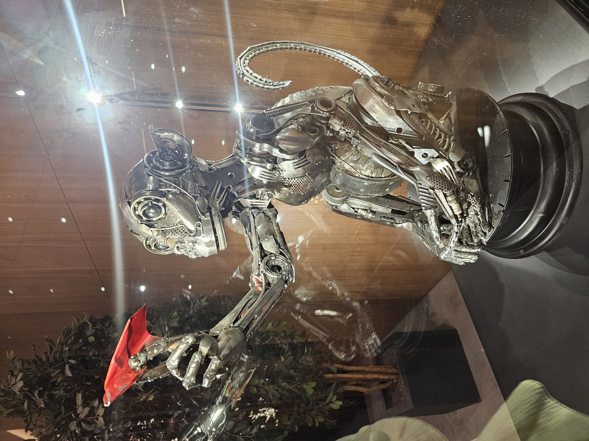 A metallic sculpture of a monkey sits inside a glass enclosure. The monkey is intricately crafted from various metal parts, resembling mechanical components. It is holding a small, bright red object in its right hand. The sculpture is seated on a dark, circular base. Behind the monkey, a large plant with green leaves is visible, potted in a dark rectangular planter. The background consists of a wooden paneled wall with subtle lighting. Reflections of lights and the surrounding environment are visible on the glass surface of the enclosure. The overall scene suggests an indoor display, possibly in a gallery or public space within Arnavutköy, Turkey.