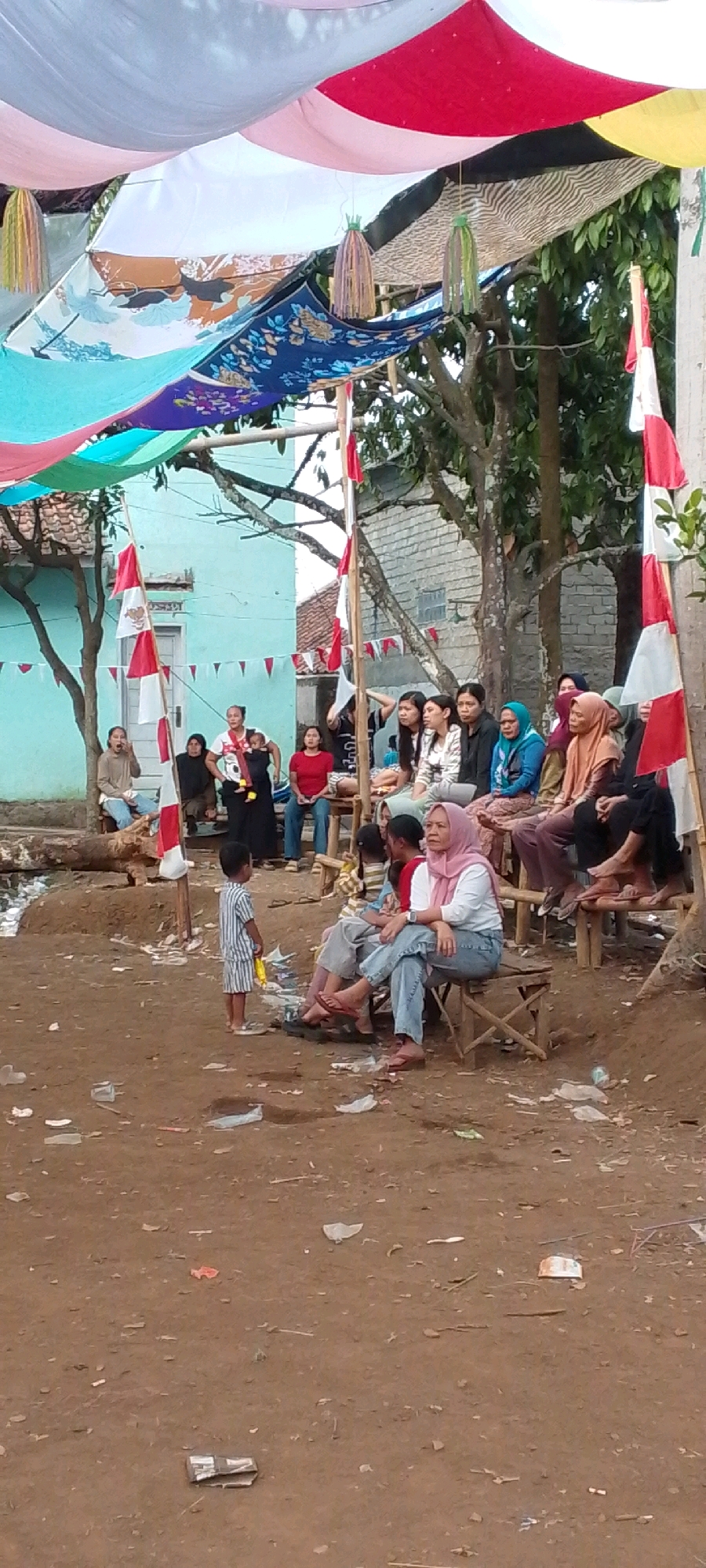 A medium shot shows a group of people, likely locals, gathered outdoors in what appears to be the city of Cilaku, Indonesia. The setting is rustic, with a dirt ground covered in scattered debris, suggesting an informal gathering or event. Above, colorful fabrics are draped to create a temporary canopy. Red and white flags, possibly Indonesian flags, are visible, attached to wooden poles.
In the foreground, a young boy in a striped outfit stands facing away from the camera. Behind him, several people are seated on wooden benches, facing a direction outside the frame. Most are women, some wearing hijabs. The group appears to be observing something or participating in an event. Some individuals are engaged in conversation, while others seem contemplative. The overall atmosphere suggests a community event or informal social gathering. The background features a building with light blue walls and a tiled roof, and trees with lush green foliage.