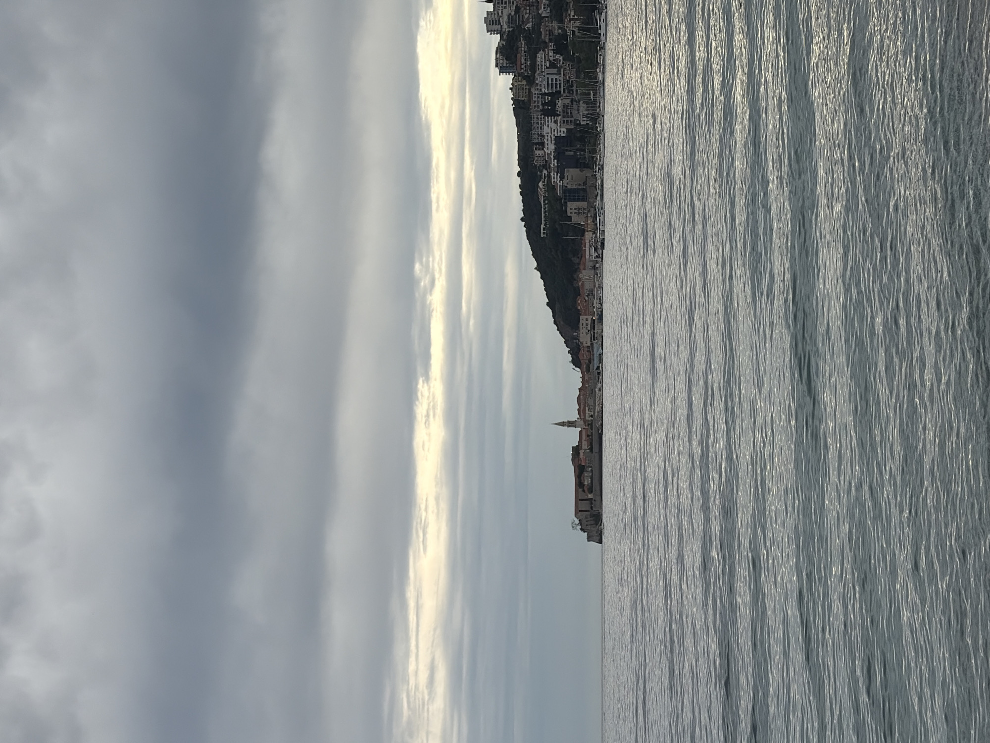 The image shows a coastal scene, likely taken from the sea looking towards the land. The water is calm with visible ripples. The sky is overcast with layered clouds. The shoreline features a city with buildings densely packed together, extending upwards along a slope. A prominent church steeple can be seen among the buildings. This is most likely the old city of Budva, Montenegro, as it features a characteristic coastline and a prominent church in its old town. There are no visible people.