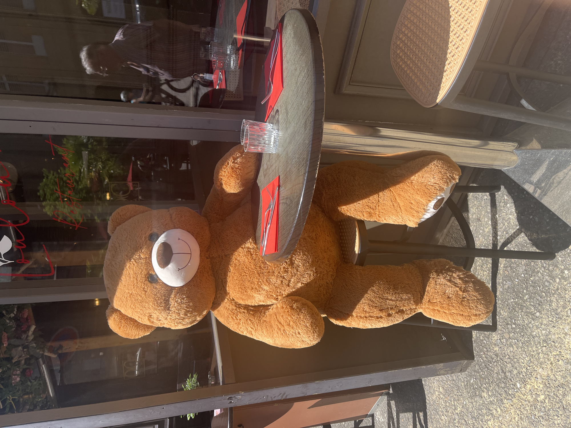 A large brown teddy bear is sitting at an outdoor cafe table. The table is set with red napkins and silverware. A glass sits on the table. The cafe is in Metz, France, identifiable by the reflection in the glass of the window and the building style.