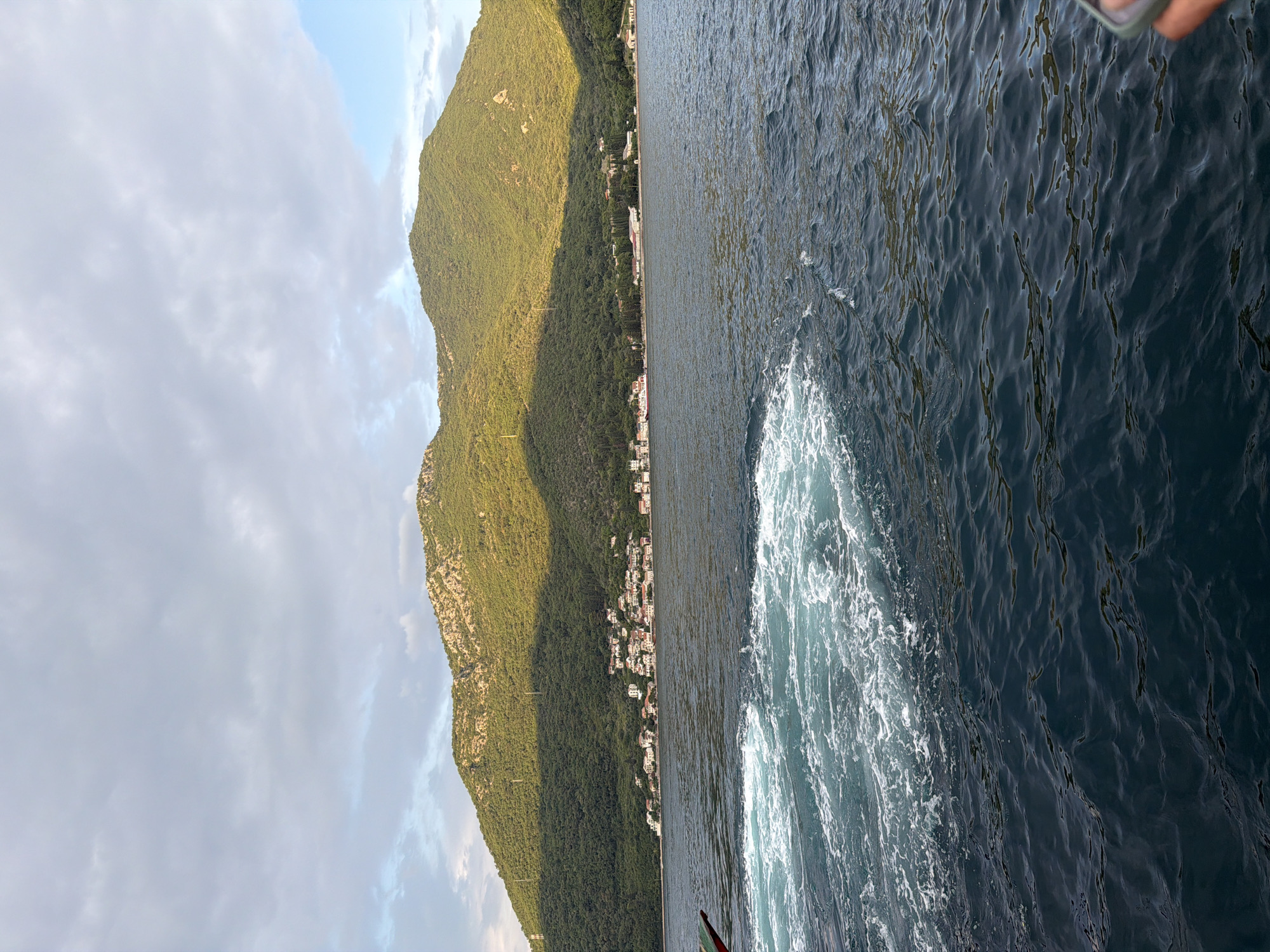 The image depicts a view of the Bay of Kotor near the village of Đurići, Montenegro. The foreground shows turbulent, foamy water, indicative of a boat's wake, with the deep blue water extending towards the shore. In the midground, a strip of coastline is visible, featuring a cluster of white buildings with red roofs, nestled against a lush green mountainside. The mountains dominate the background, with their slopes covered in dense vegetation and rocky outcrops. Sunlight casts a warm glow on the upper portions of the mountains, creating a contrast with the shadowed valleys. The sky above is partly cloudy, with patches of blue sky visible amongst the grey clouds. A hand holding a phone is partially visible in the lower right corner of the frame.