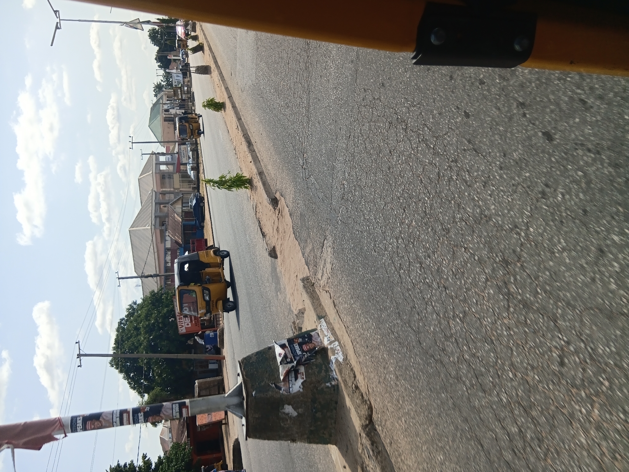 The image depicts a street scene in Jalingo, Nigeria, with a focus on a yellow auto-rickshaw (Keke Napep) driving on the asphalt road. The scene is captured from a moving vehicle, likely another Keke Napep, as indicated by the partial view of a yellow frame on the right side of the image. The road is paved and shows some signs of wear and tear, with visible cracks and patches. On the left side of the road, a concrete structure, possibly a utility pole base or a public fixture, is covered in torn posters and debris. A tall metal pole with banners and election-related posters is also prominent on the left. Several yellow auto-rickshaws are visible on the road, with one prominently featured in the middle ground. In the background, there are buildings, including shops and residential structures, with corrugated iron roofs. Trees and utility poles with overhead wires are also part of the urban landscape. The sky is partly cloudy with scattered white clouds against a blue background. The overall scene suggests a typical day on a busy street in Jalingo, with vehicular traffic and commercial activity.