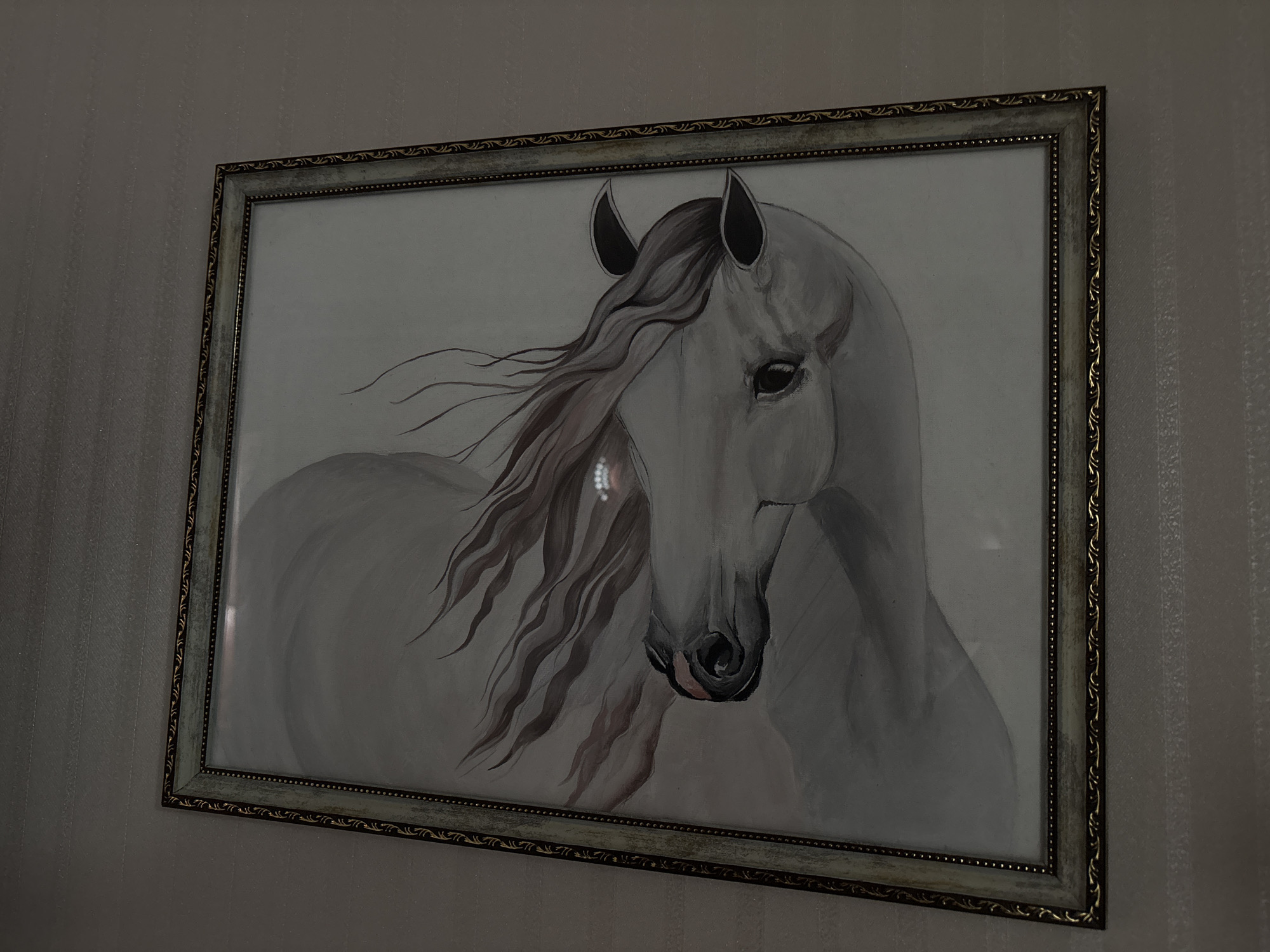 A framed painting of a white horse is hanging on a striped wallpapered wall. The horse is depicted in profile, facing left, with its mane flowing. The painting is in grayscale with subtle hints of brown in the horse's mane. The frame is ornate, with a light-colored outer edge and a darker, patterned inner trim. The lighting suggests an indoor environment. The location is Shepetivka, Ukraine.