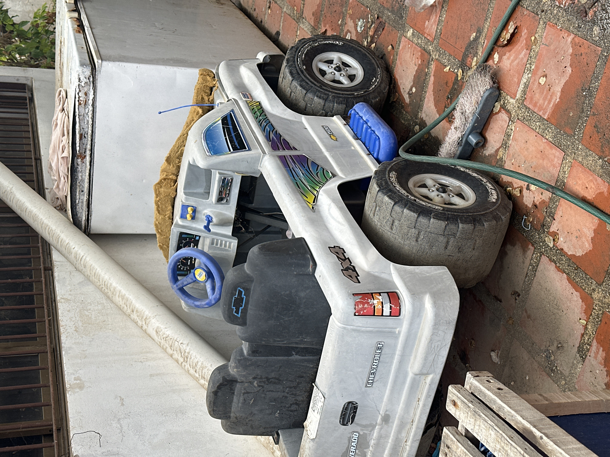 This image captures a weathered, white Chevrolet Silverado 4x4 toy truck positioned outdoors on a reddish-brown tiled surface. The truck, likely a child's ride-on vehicle, shows signs of extensive use and exposure to the elements, with scuffs, dirt, and fading.
Key features of the toy truck include:
* **Color and Decals:** Predominantly white, with a colorful, rainbow-like graphic decal on the side of the cab. Visible text includes "CHEVROLET," "SILVERADO," "4X4," and "1500." A blue Chevrolet logo is on the front fender/bumper area.
* **Wheels:** Four large, black plastic wheels with silver hubs, mimicking off-road tires.
* **Interior:** A blue steering wheel with a Chevrolet logo is visible, along with a simple dashboard featuring gauges and buttons. An antenna protrudes from the cab.
* **Accessories:** A blue plastic container, possibly a prop gas can, is attached to the side of the truck bed. A yellowish, soft material (perhaps foam) is tucked around the driver's seat area.
The scene appears to be a back patio or side yard. The ground is covered in red terracotta or brick-like tiles, which show patches of dampness, stains, and debris like small leaves.
Other objects in the environment include:
* **Hose and Brush:** A green garden hose is coiled on the ground near a black and white scrub brush.
* **Structures:** A large, white, weathered appliance (possibly an old refrigerator or freezer) with visible rust and peeling paint is positioned behind the truck. A long, white pipe runs horizontally along a wall in the background. Part of a metal grille or fence is also visible.
* **Miscellaneous:** A wooden pallet or crate is partially visible in the lower right corner. Some greenery from plants can be seen in the upper left.
The lighting suggests it is daytime, and the overall impression is one of a storage area for outdoor items, possibly in a state of disuse or disrepair, in a warm, likely humid climate given the location context of Juan Griego, Venezuela. No people are visible.