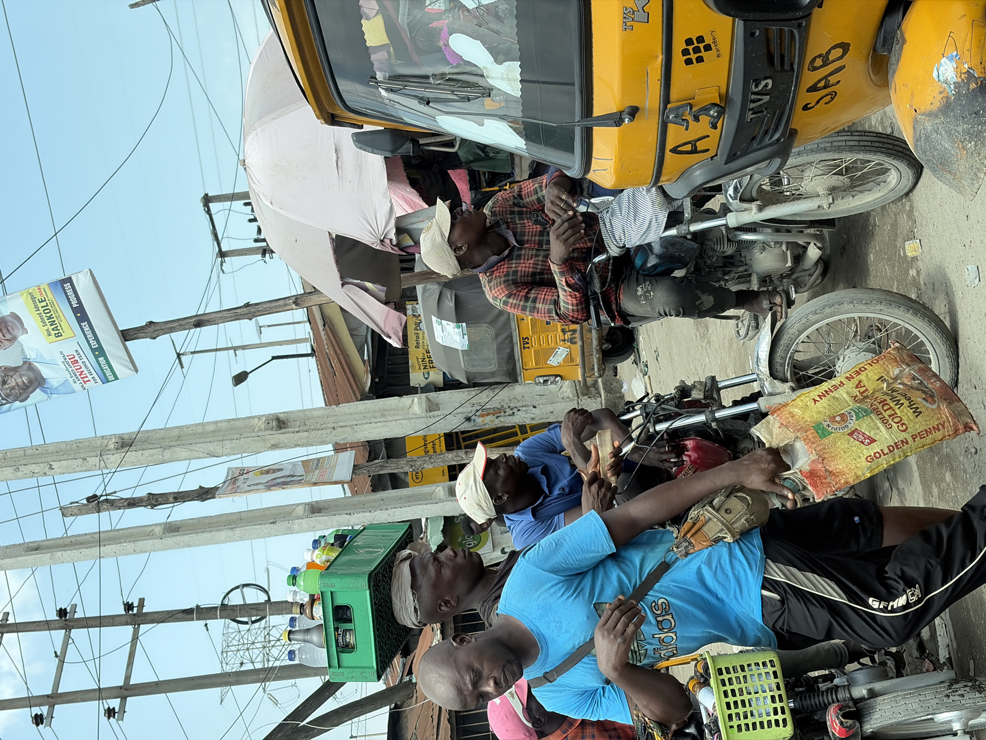 The image captures a street scene, likely in Lagos, Nigeria, featuring men on motorcycles amidst an urban environment.
In the foreground, three men are positioned on their motorcycles. The man closest to the camera wears a blue Adidas t-shirt, black and white track pants, and a cross-body bag. A green crate filled with water bottles is attached to his bike. Another man, wearing a blue shirt and a white cap, is seated behind him. He is handling what looks like a piece of wood. A bag labeled "GOLDEN PENNY" lies across his lap. Further back, a third man in a plaid shirt and a white cap is on a yellow auto rickshaw, known locally as a "keke," labeled "TVS" and "SAB".
Overhead, there are power lines and utility poles with political campaign posters advocating for Bola Ahmed Tinubu and Saheed Adesegun Bankole. One poster features a photo of Bola Ahmed Tinubu and encourages voters to vote for his second term.
In the background, buildings and structures characteristic of an urban environment are visible. A light-colored awning or umbrella provides shade nearby.