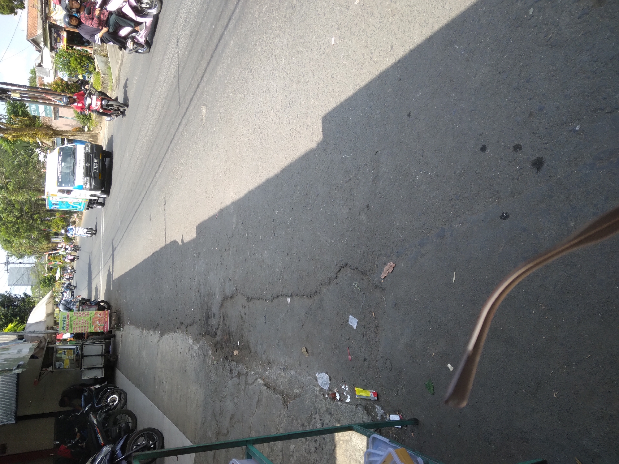 The image depicts a street scene in Sokaraja, Indonesia, likely during daylight hours. On the left side of the street, several parked motorcycles are visible in front of a building with an awning. A food stall with a sign that reads "Pentol Wara Wiri" is also present. The street itself appears to be paved and shows signs of wear and tear, with some cracks and debris scattered on the surface. A shadow of a building or trees falls across the road. Several people are seen on motorcycles, some riding individually and others in pairs, navigating the street. A white delivery truck is also present on the road. The environment appears to be an urban or semi-urban setting, with trees and buildings lining the street. A pair of eyeglasses is prominently in the foreground, partially obscuring the view of the street.