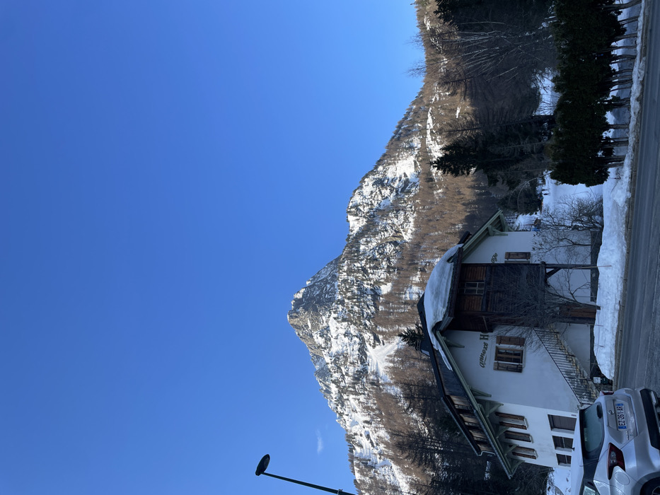 This image depicts a winter scene in Vallorcine, France, under a clear, bright blue sky.
In the foreground, a paved road, partially clear of snow, spans the lower portion of the frame. On the left, a silver Nissan Micra, bearing license plate EZ-261-AR, is parked facing away from the viewer. A tall, black streetlamp is positioned to the far left. A line of trimmed evergreen bushes, partially covered in snow, runs along the right side of the road.
The midground features a multi-story building with a white facade and dark brown wooden upper sections, labeled "Annexe H" with additional faded text. Snow covers parts of its gabled roof. An external metal staircase with white risers provides access to the upper level. Bare trees and scattered evergreens surround the building and extend towards the background. On a snowy slope behind the bushes, two distant figures are discernible, suggesting winter activity.
The background is dominated by a rugged, snow-covered mountain, featuring rocky peaks and slopes interspersed with coniferous trees and bare deciduous trees. Extensive snow blankets the mountain, and patches of darker rock are visible.