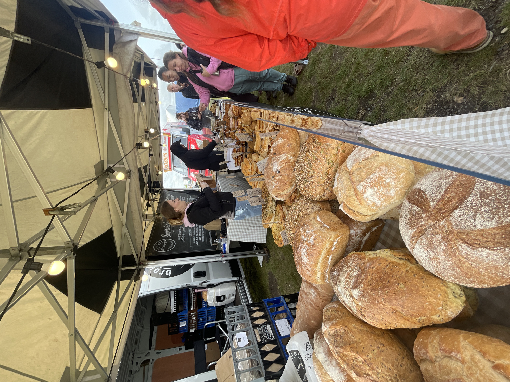 At a market stall in Sevenoaks, United Kingdom, a baker sells bread to customers. The stall is covered by a white tent with strung lights. A white van displaying "bro" is parked nearby. A variety of bread loaves are displayed on a checkered cloth. Several customers are at the stall, including one wearing a pink top and another in a red jacket.