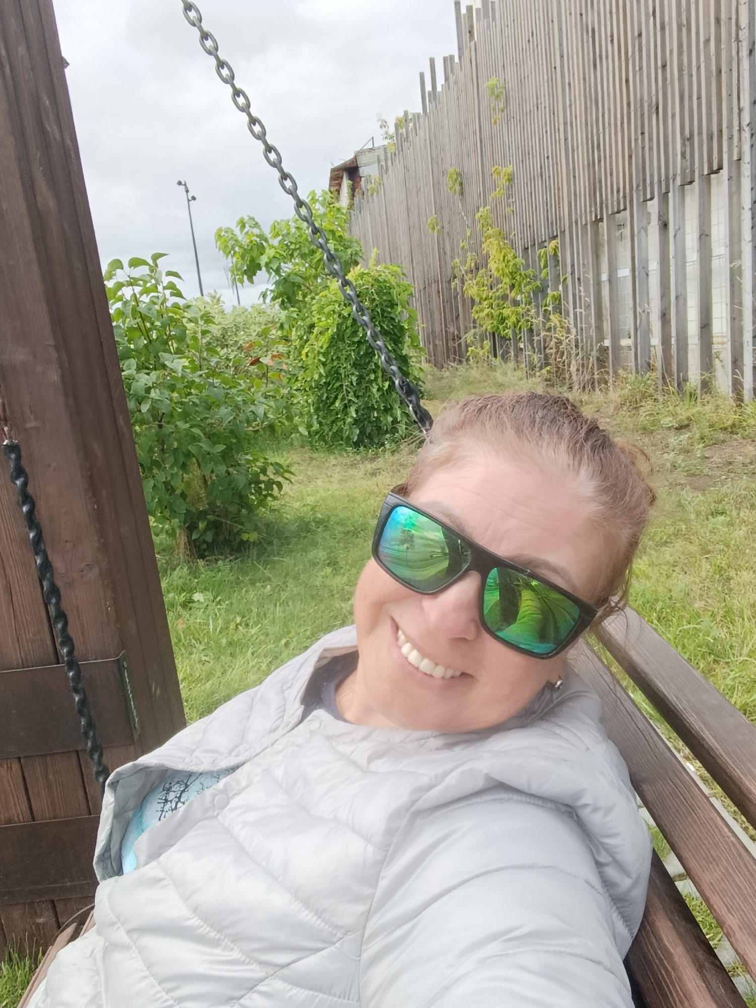 A woman with light brown hair, wearing sunglasses and a light gray puffer jacket, sits on a wooden swing. She is smiling and looking towards the camera. Her sunglasses reflect greenery. In the background, there is a wooden fence, lush green bushes, and a cloudy sky. The scene appears to be outdoors in a park or garden, possibly in the city of Engels, Russia.