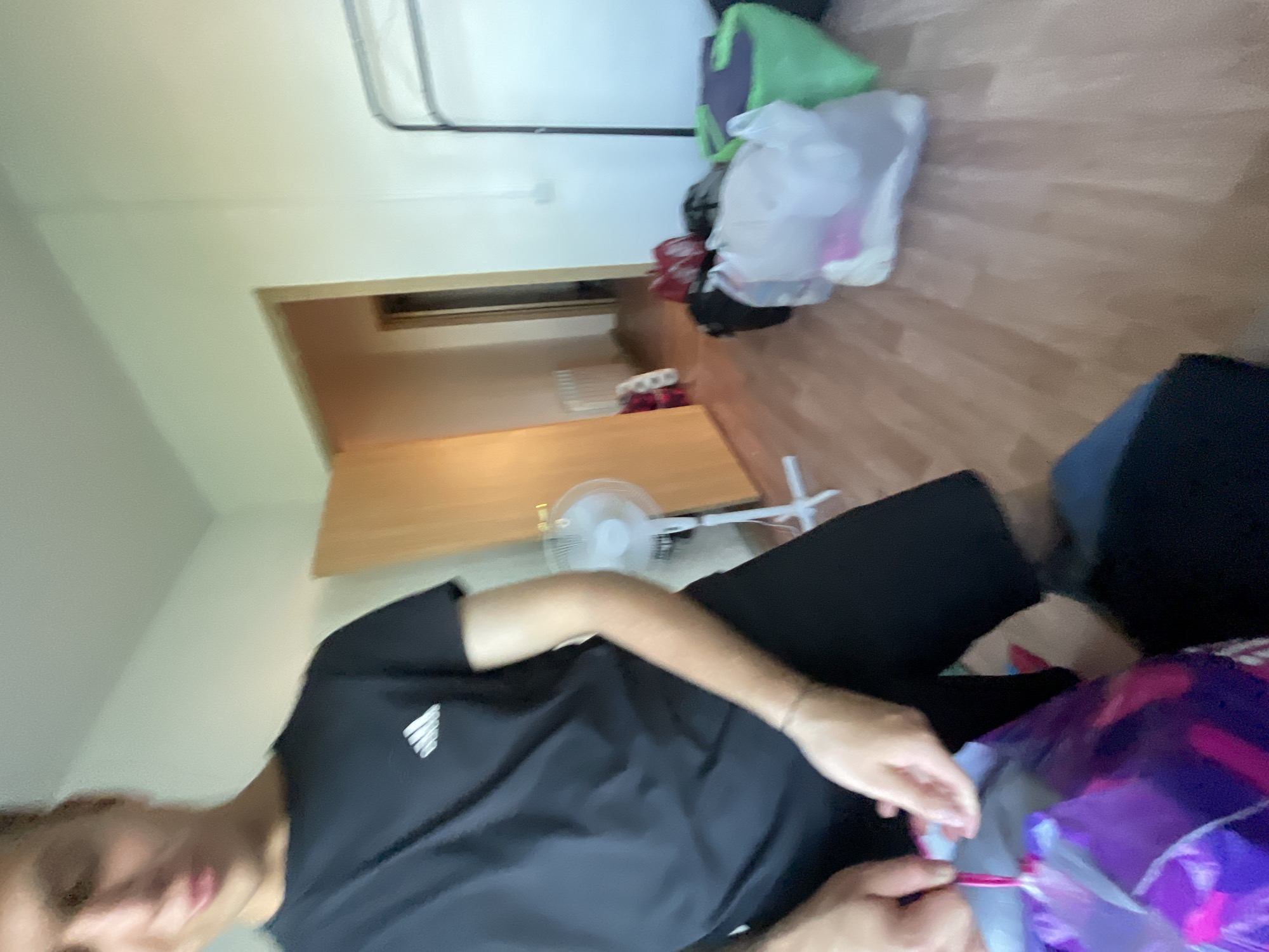 A person wearing a black Adidas t-shirt and black shorts is holding a purple shopping bag in their hands. The person appears to be in an indoor setting, possibly a room, with a wooden floor and a white wall. There is a white standing fan in the background, as well as several other bags of various colors, including red, green, and white. A doorway leading to another room or hallway is visible on the left side of the image. The scene appears to be in the city of Krasnye Baki, Russia, as indicated by the context. The overall lighting suggests it is daytime.