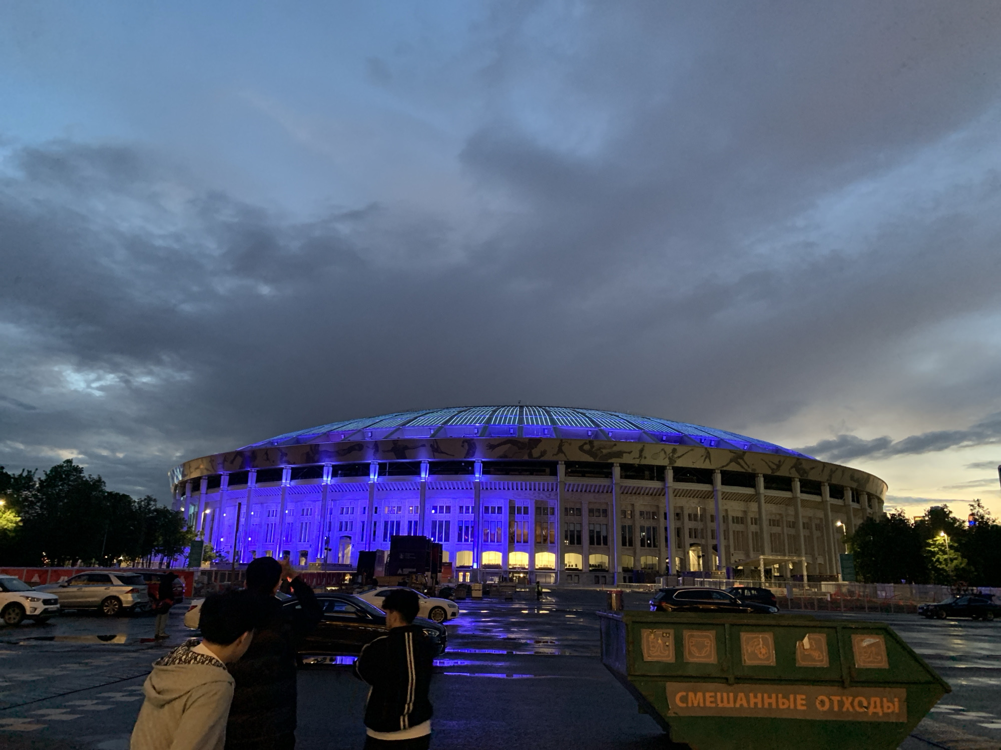 The image shows the Luzhniki Stadium in Moscow at dusk. The stadium is illuminated with blue lights. In the foreground, there is a green dumpster with the words "Смешанные отходы" written on it, which translates to "Mixed Waste". Several cars are parked nearby, and a group of people is standing in front of the cars. The sky is filled with dark, cloudy formations. The ground appears wet.