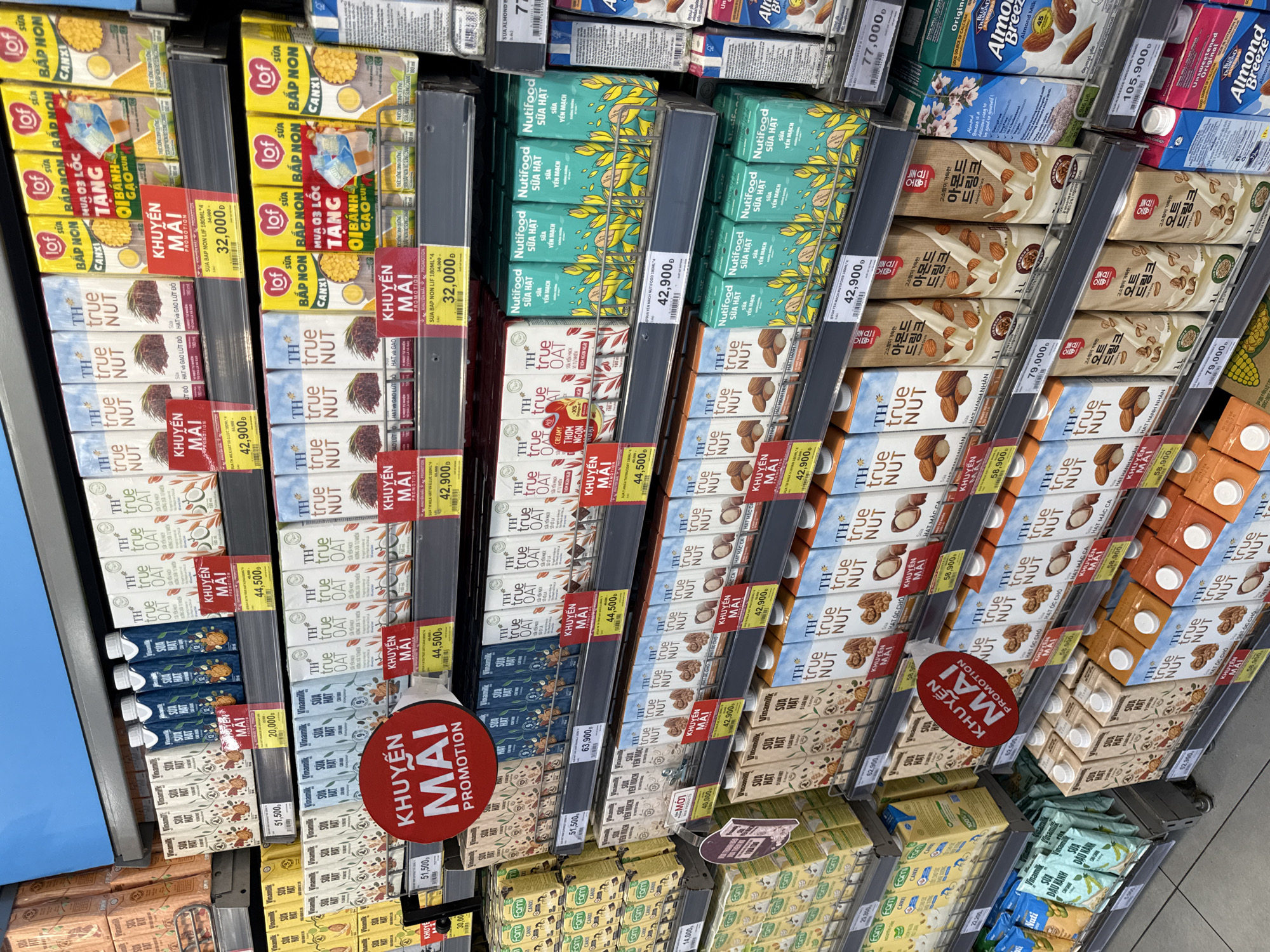 The image shows a shelf in a supermarket in Nha Trang, Vietnam, displaying various milk cartons and beverages. The products are arranged on multiple shelves, with many cartons featuring Vietnamese text and promotional labels like "KHUYEN MAI PROMOTION". The products include brands like TH true NUT, Vinamilk, Lof, Nutrifood, and Almond Breeze. Some shelves have price tags visible, indicating prices in Vietnamese Dong. The overall environment appears to be a well-stocked retail setting.