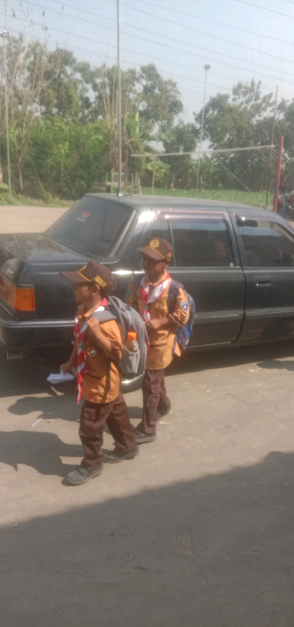 Two young boys dressed in scout uniforms are walking beside a black car. Both boys are wearing brown hats, orange scout shirts, brown pants, and dark shoes. They both have backpacks on. The boy in the foreground is facing left and holding a white paper in his left hand. The boy in the background is facing forward and slightly to the right. The car is parked on a paved surface, likely a road or parking lot, with a dirt or gravel area to the right. In the background, there are trees and what appears to be a volleyball net. The sky is light blue with some faint power lines visible. The overall scene suggests an outdoor activity or travel.