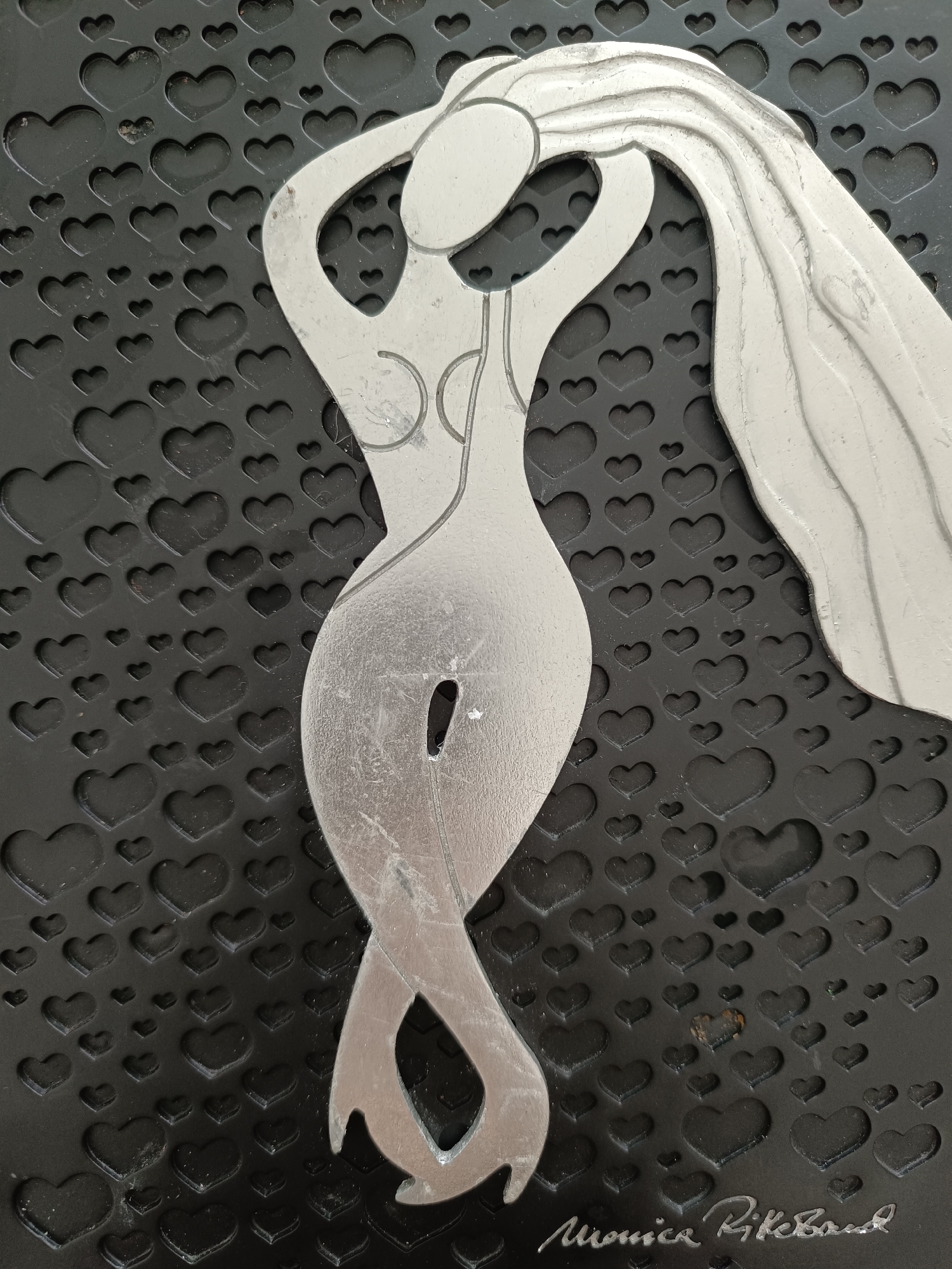A silver cutout of a woman with flowing hair and a curvaceous body is positioned on a black background textured with numerous small hearts. The woman's figure is depicted in profile, with her arms raised above her head. Her lower body transitions into two intertwined, ribbon-like legs. A small, oval-shaped opening is visible on her torso. The artwork is signed in the bottom right corner with the name "Monica Rikbord" in cursive silver lettering. The background is dark and appears to be a textured mat or surface, with the heart shapes creating a repeating pattern. The lighting casts subtle shadows, highlighting the metallic sheen of the cutout and the depth of the background texture. There is no discernible location in the city of Hovden, Norway, associated with this image.