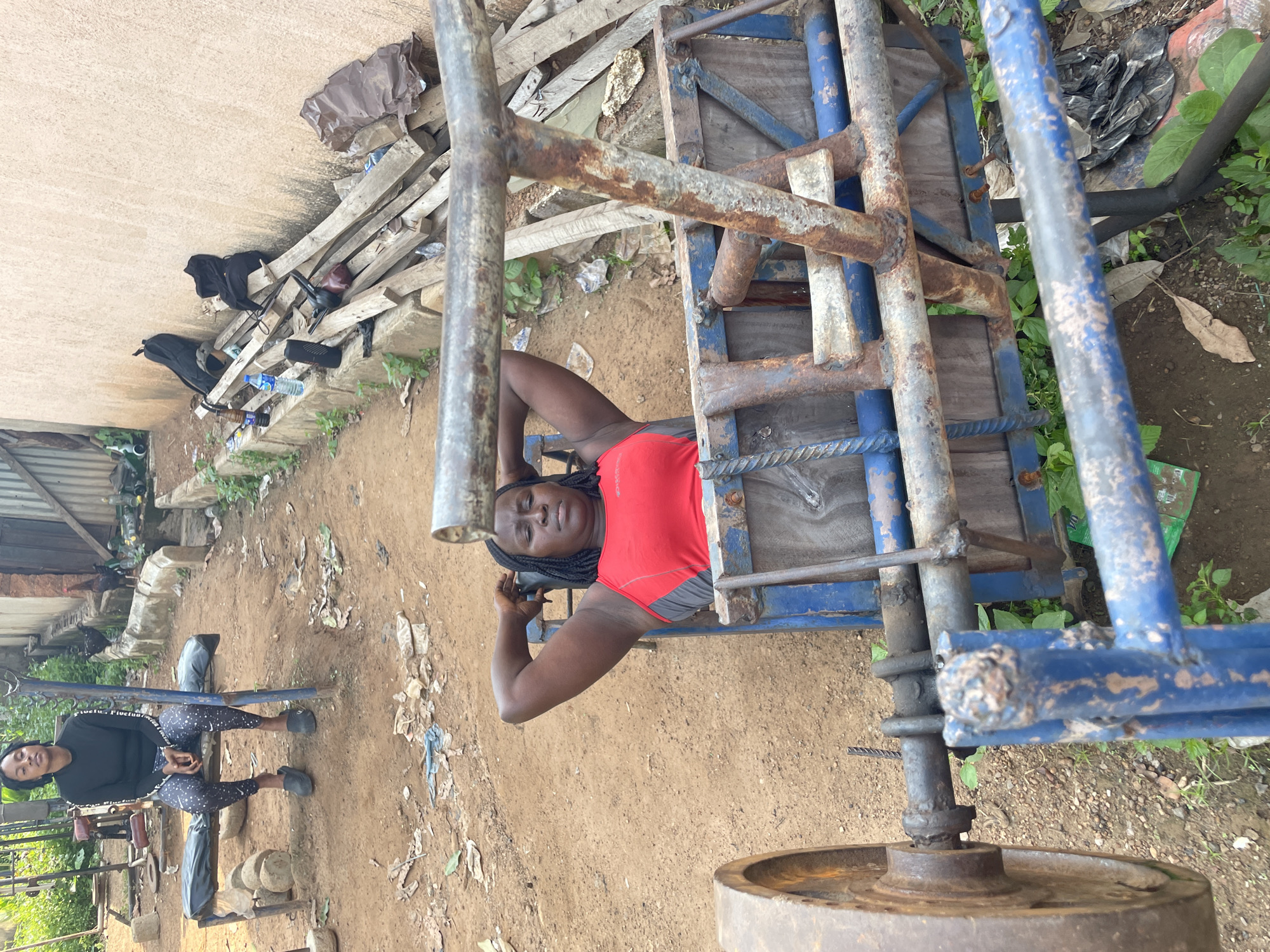 This media file depicts an outdoor scene in Akwanga, Nigeria. Two individuals are present, both appearing to be women. One woman is lying down on a makeshift weightlifting apparatus constructed from scrap metal and wood. She is wearing a red sports top and appears to be engaged in some form of exercise, possibly a chest press, with her arms extended above her. The other woman is seated on a bench nearby, wearing a black long-sleeved shirt and patterned leggings, also seemingly involved in a workout.
The setting is rustic and appears to be an informal outdoor gym or training area. The ground is sandy dirt, littered with debris including pieces of wood, plastic bottles, and other discarded materials. Various metal components, including pipes and what seem to be weights, are scattered around, suggesting a DIY approach to fitness equipment. In the background, a building with a textured wall is visible, as well as some vegetation and more discarded items. The overall impression is one of a resourceful and perhaps impoverished environment where individuals are making do with available materials for physical training. The lighting suggests it is daytime, and the weather appears to be fair. The subjects' expressions are neutral, focused on their activities. There is no visible text in the image.
