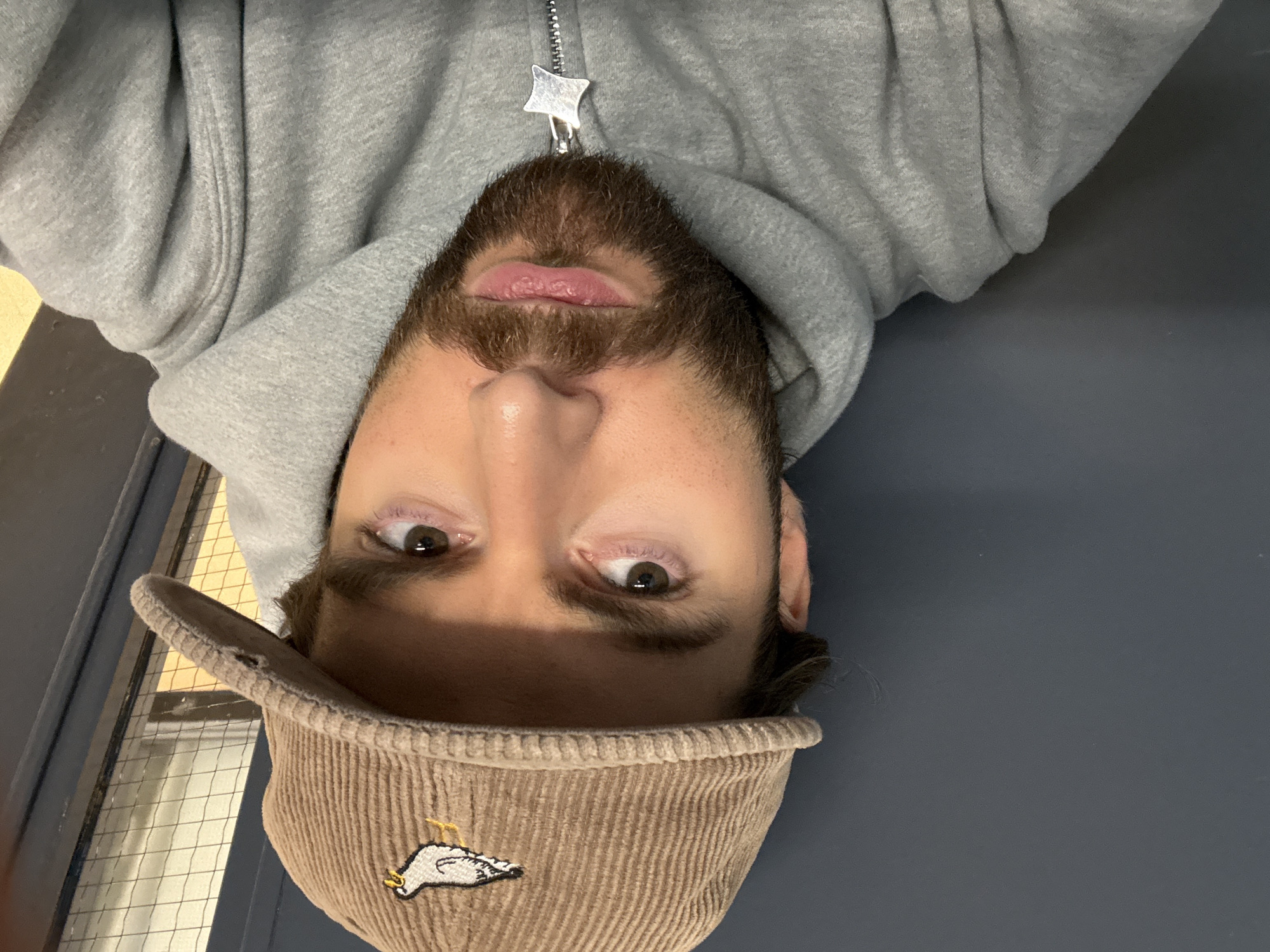 A man is shown from a low angle, wearing a grey hooded sweatshirt with a star-shaped zipper pull and a brown corduroy baseball cap with a white bird logo. He has brown eyes, a beard, and light skin. The background appears to be a grey-blue wall on the right and a window with vertical metal bars on the left, suggesting an institutional setting, possibly in Montreal, Canada. The photograph is rotated ninety degrees.