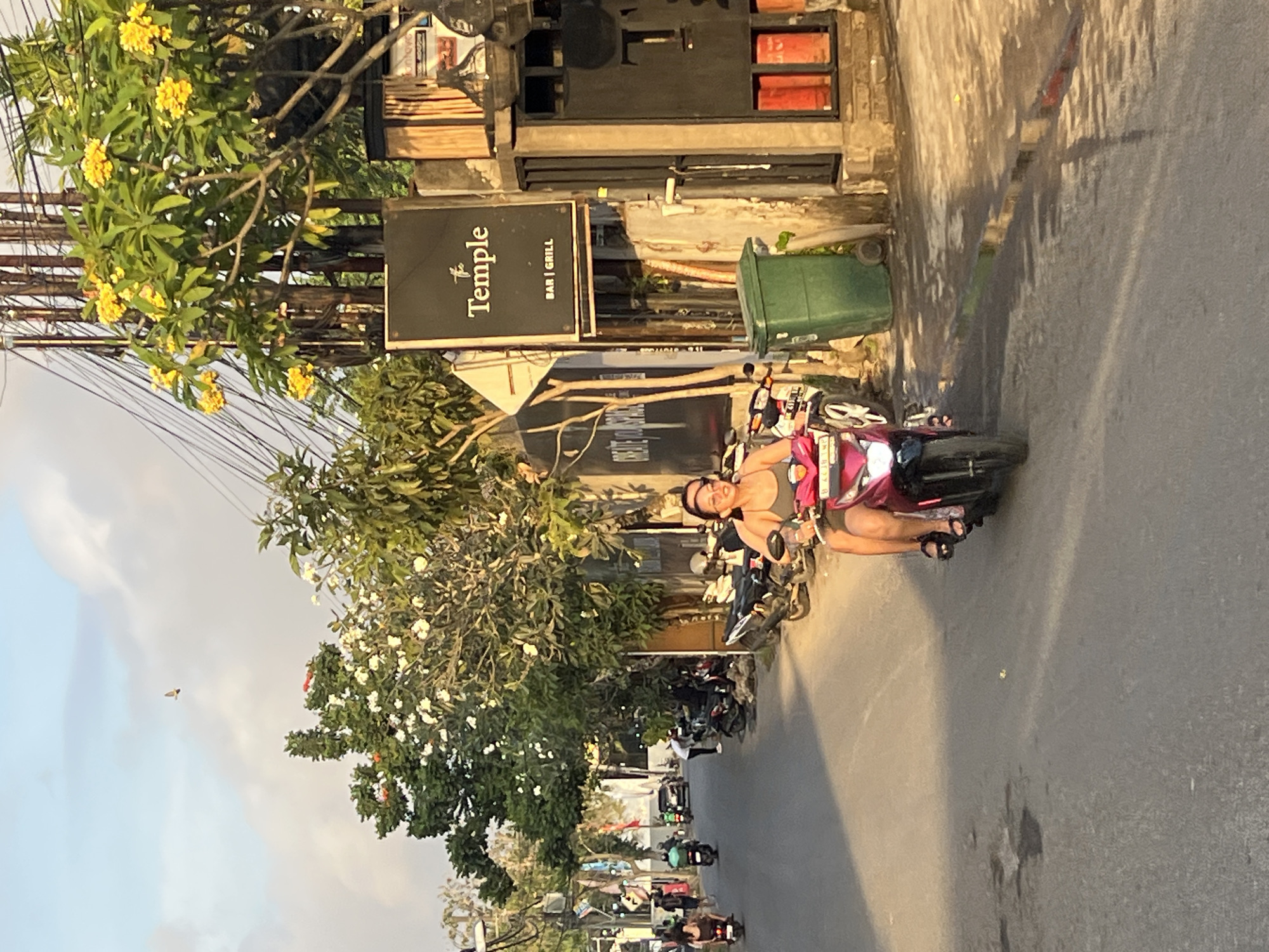 An outdoor, eye-level, medium shot shows a woman on a pink scooter on a street in Kuta Selatan, Indonesia. The woman is wearing a grey tank top and shorts and sunglasses. She is smiling and looking ahead. The scooter is moving towards the viewer. In the background, there are trees, buildings, and other scooters. The sun is setting, casting a warm glow on the scene. A bird is flying in the sky.